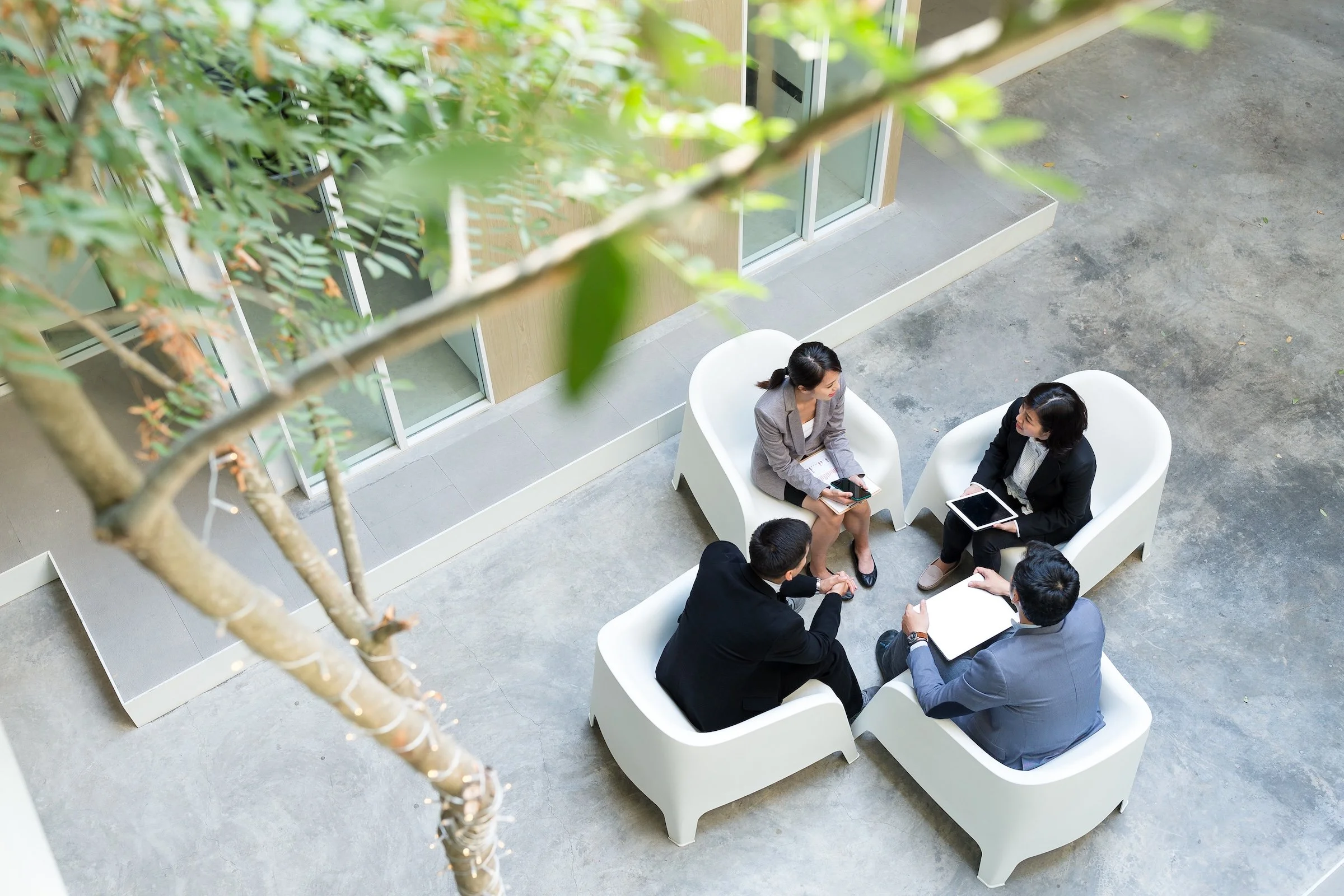 Four people engaged in a conversation sitting on white modern chairs outdoors near a building with large windows, partially obscured by tree branches.