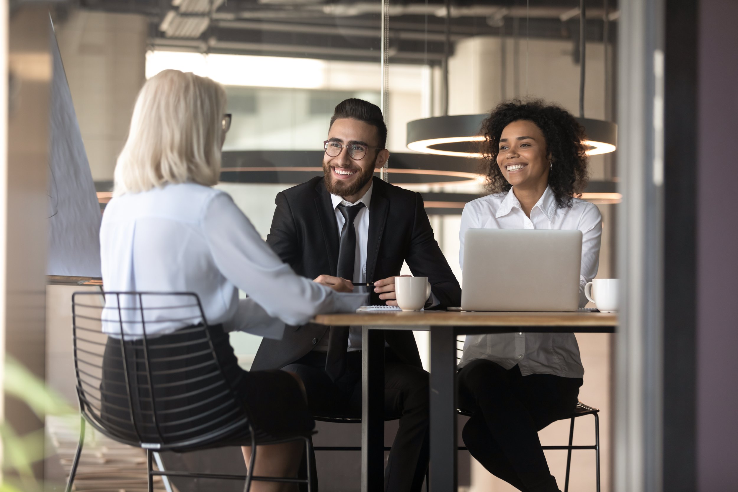 Three professionals, two women and one man, sitting at a round table in a modern office space engaged in a conversation. The woman on the left has blonde hair, the man in the center has dark hair and a beard, and the woman on the right has curly dark hair. They are smiling and appear to be discussing work, with laptops and coffee mugs on the table.