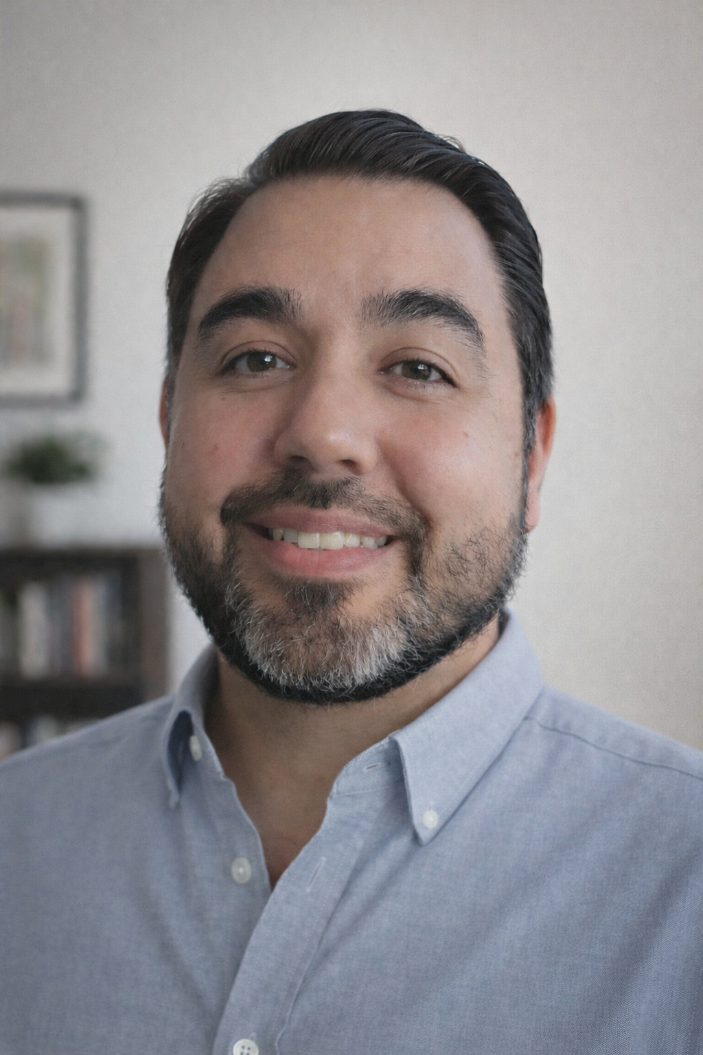 Close-up of smiling man with dark hair, beard, and light blue button-up shirt, indoors with blurred background.