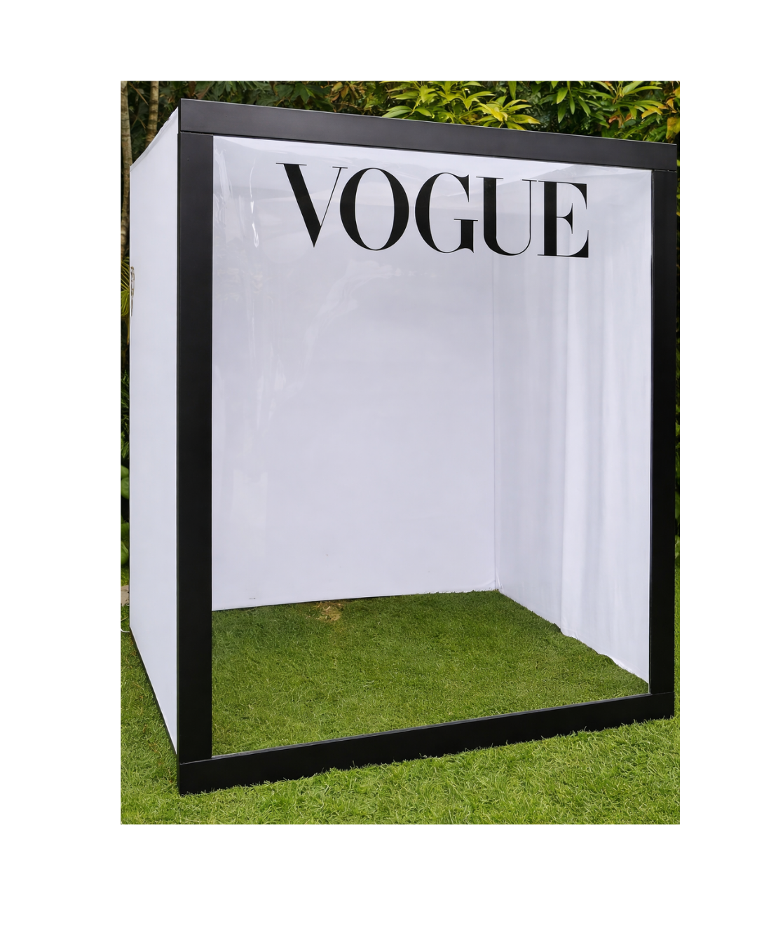 Empty photo booth with black frame, white backdrop, and the word 'VOGUE' at the top, set on grass outdoors.