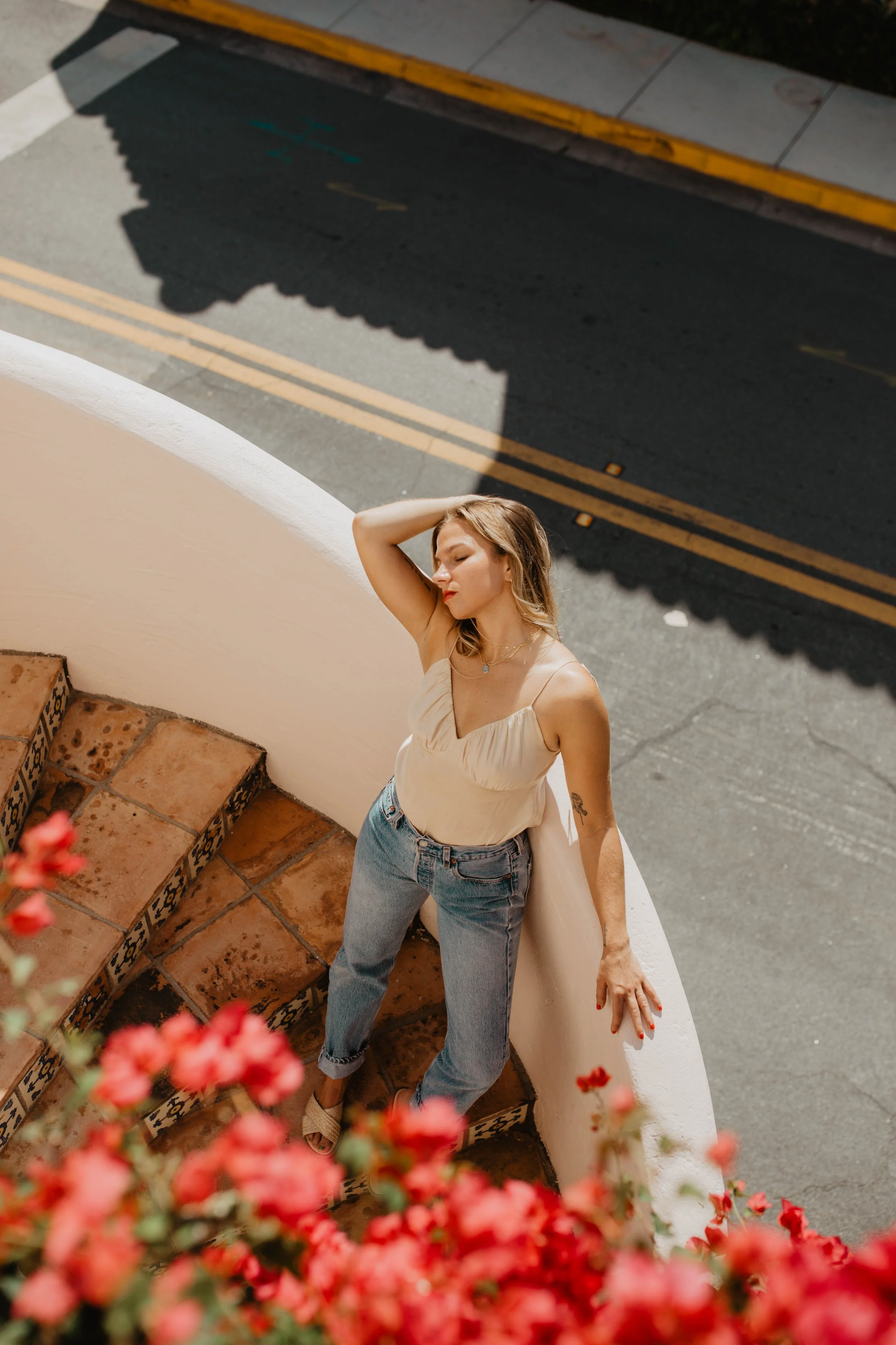 A woman with blonde hair wearing a beige top and blue jeans, standing on a curved staircase with ornate tile trim, overlooking a street with yellow lines, and flowers in the foreground.