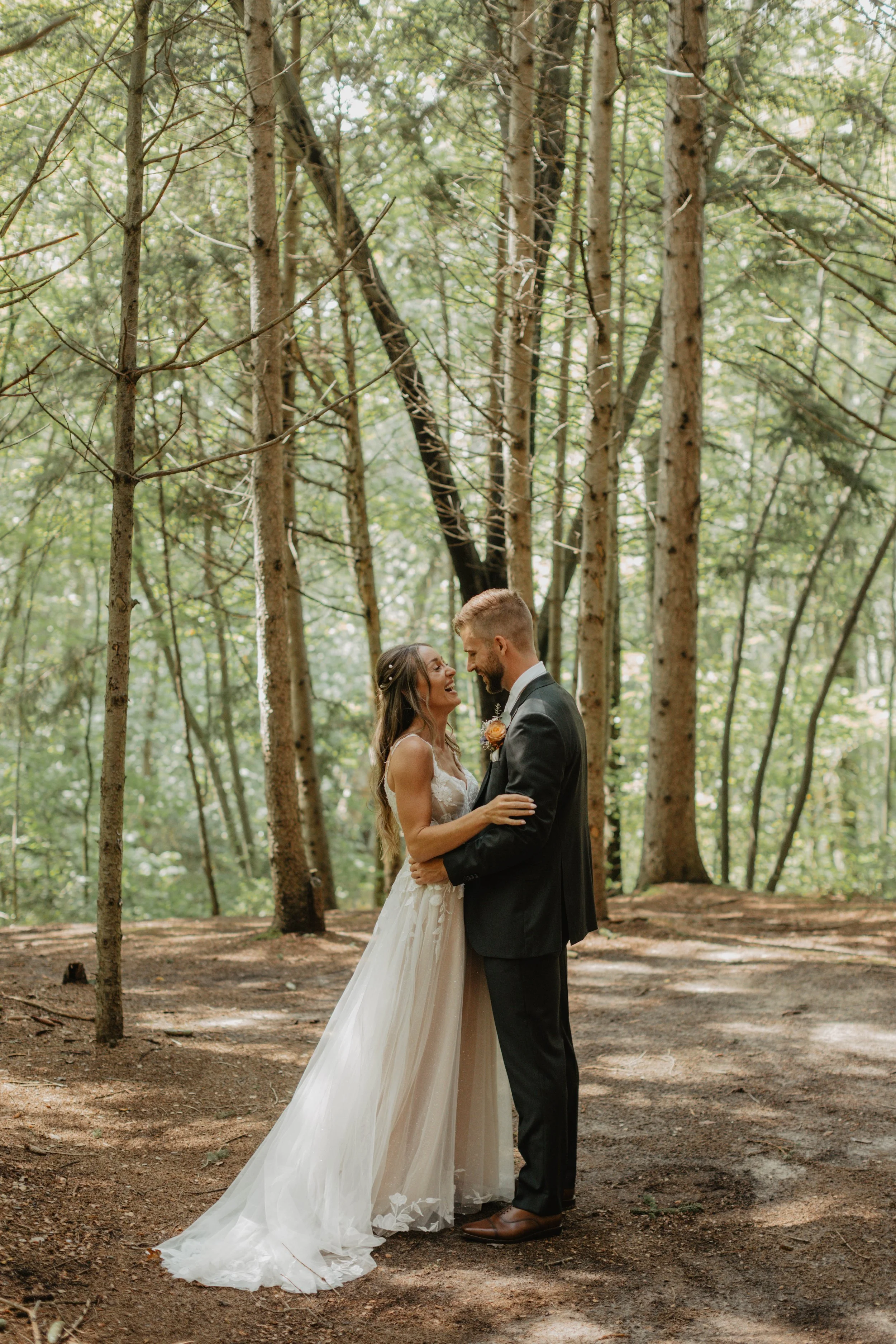 A bride and groom are embracing and smiling at each other in a forest with tall trees and green foliage.