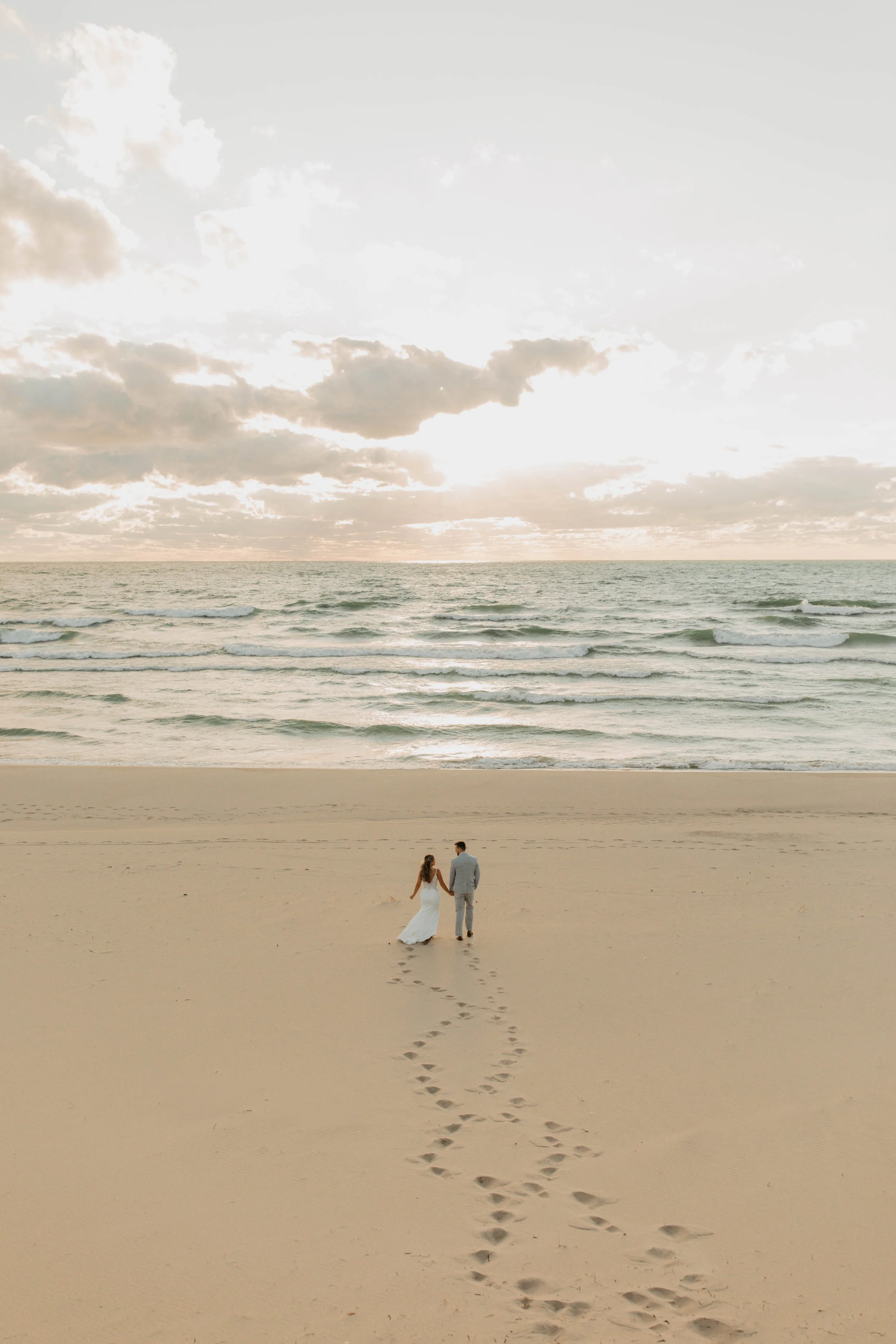 A couple dressed in wedding attire walking hand in hand along a sandy beach during sunset, with footprints trailing behind them, and the ocean waves in the background.
