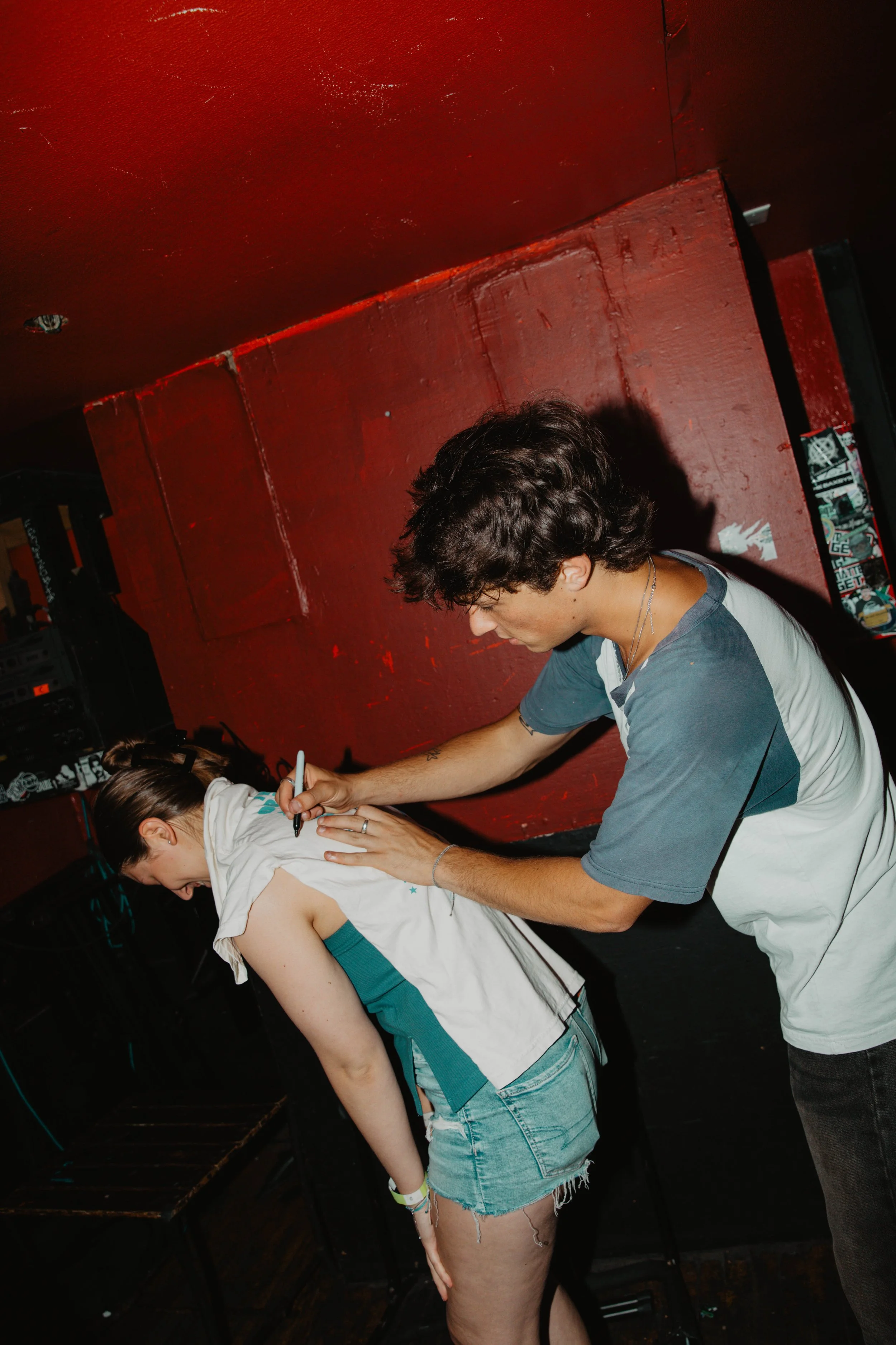 Kevian Kramer signs the back of a young woman's shirt in a dimly lit room with red walls, after a concert.