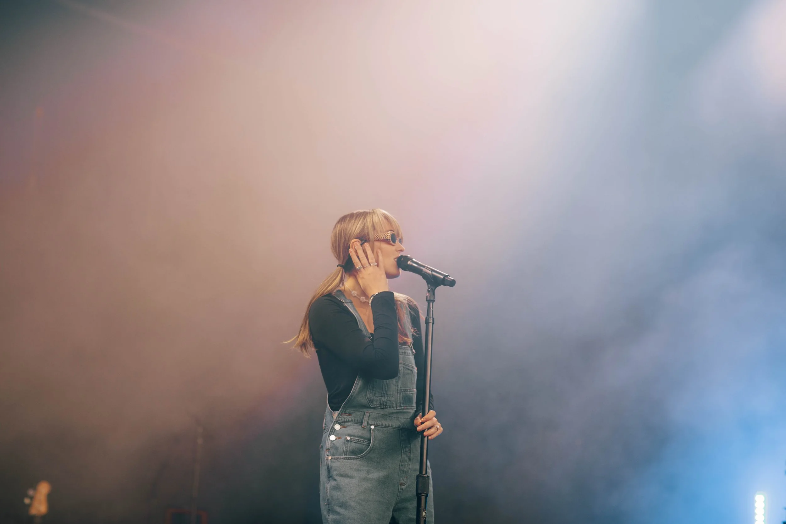 Sasha Sloan  is performing on stage, wearing sunglasses, with one hand near her face and the other holding a microphone, performing in Detroit, Michigan.