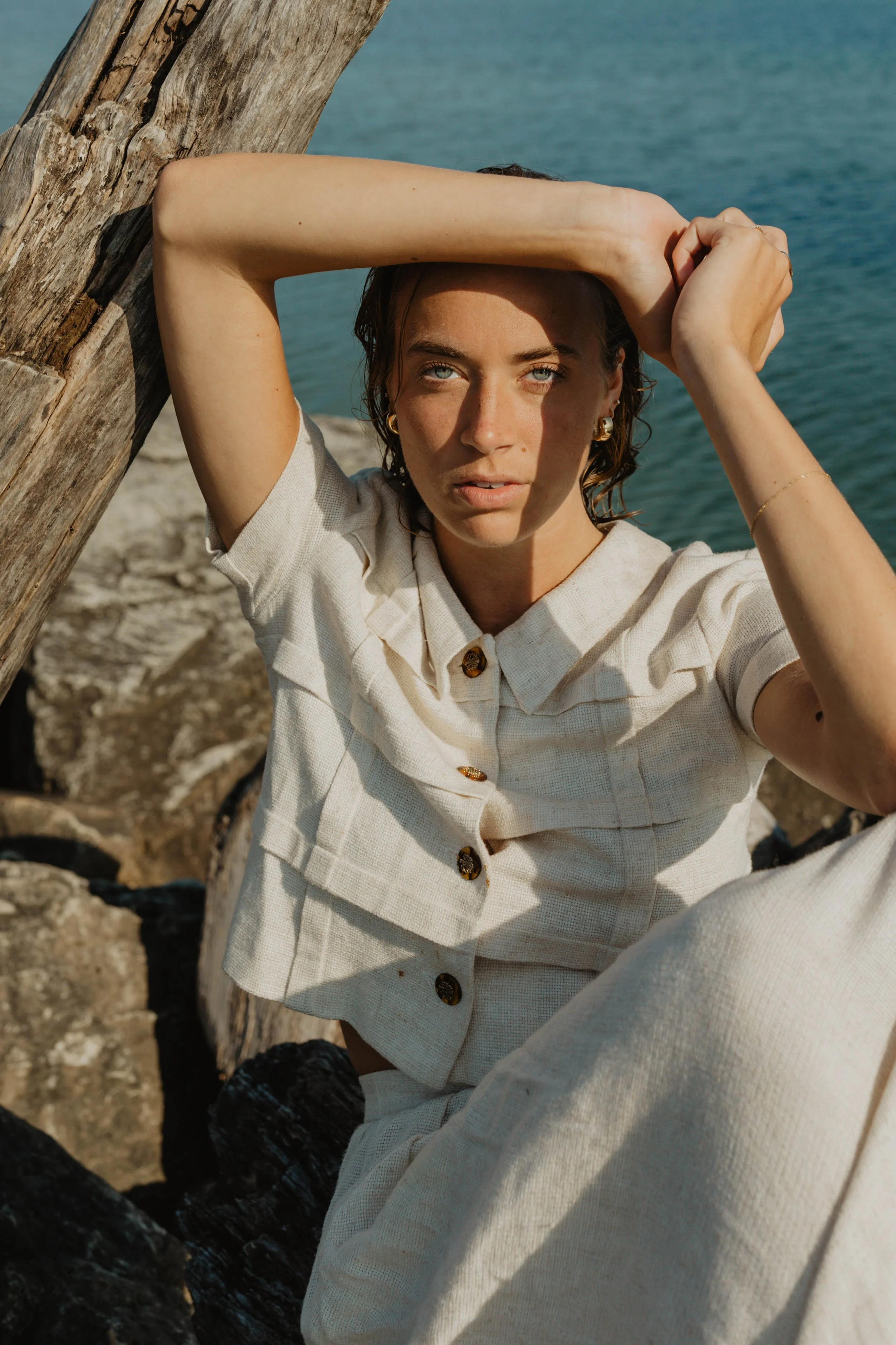A woman with short, wavy hair and blue eyes sitting by the water, with her arm resting on a wooden log, wearing a light-colored short-sleeved shirt and matching pants, surrounded by rocks.