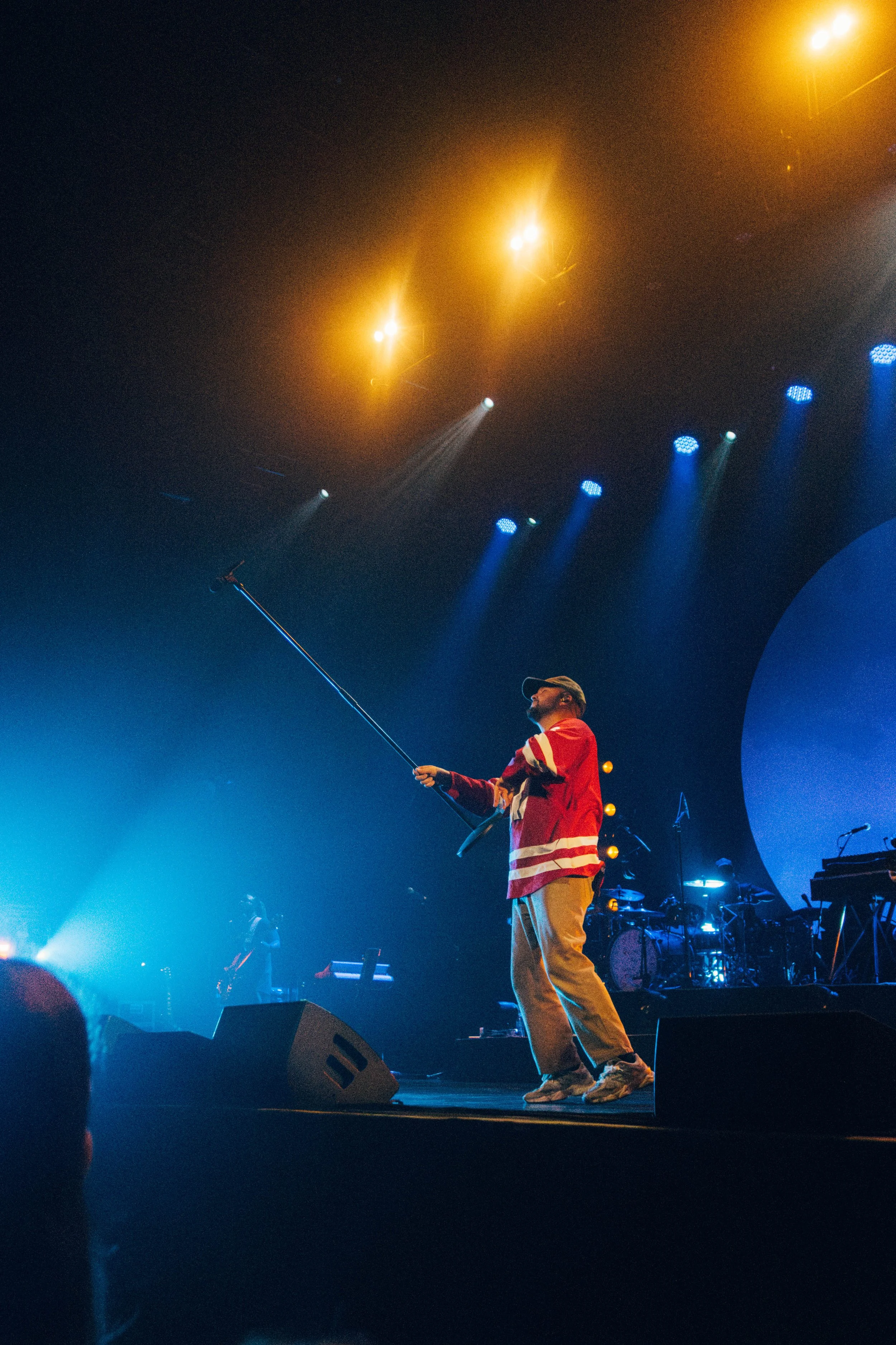 The music artist Quinn XCII performing on stage with a microphone stand, wearing a red and white hockey jersey, beige pants, and a cap, under blue and yellow stage lights.