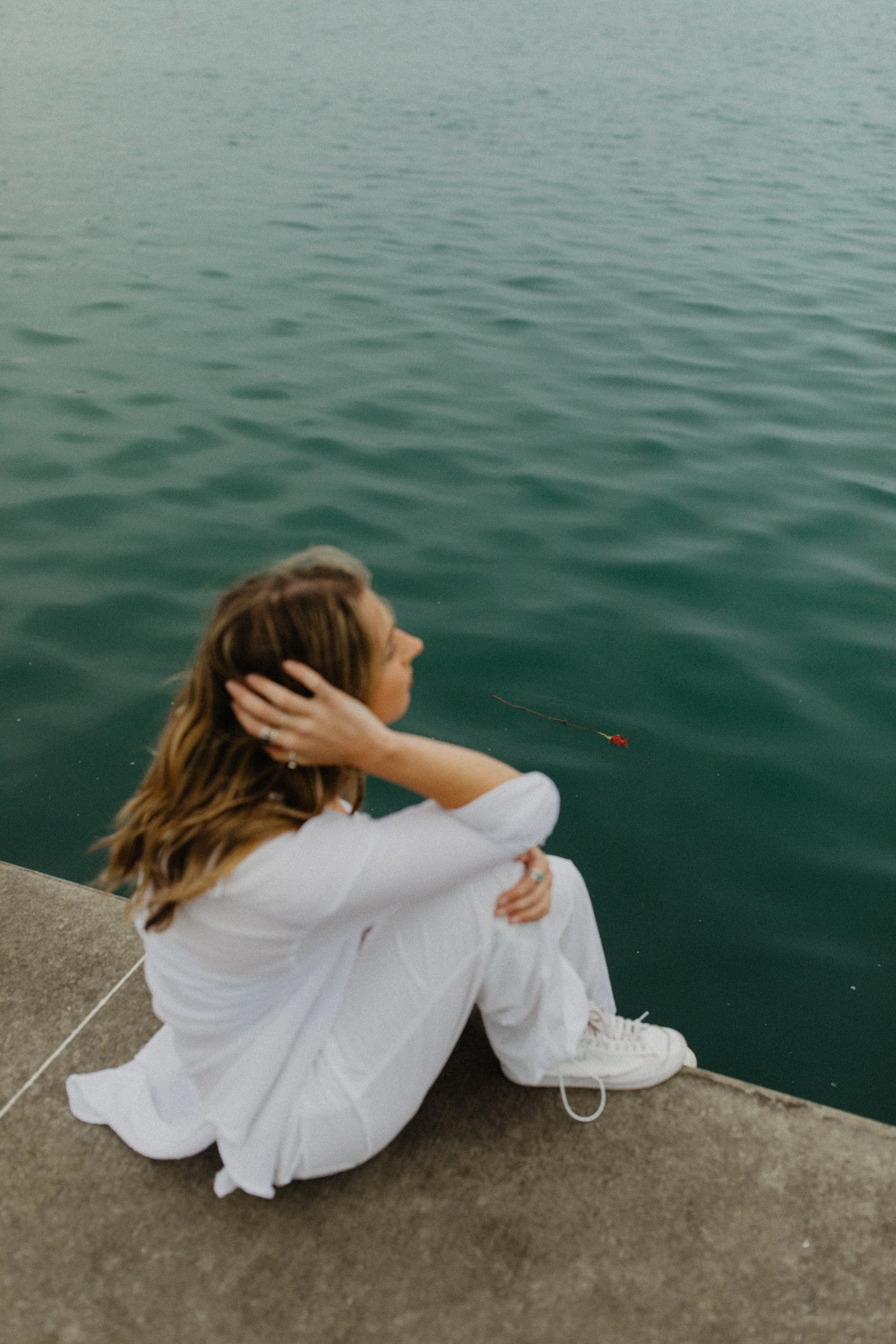 Woman in white sitting on concrete dock with her legs crossed, gazing at water with a single red flower floating nearby.