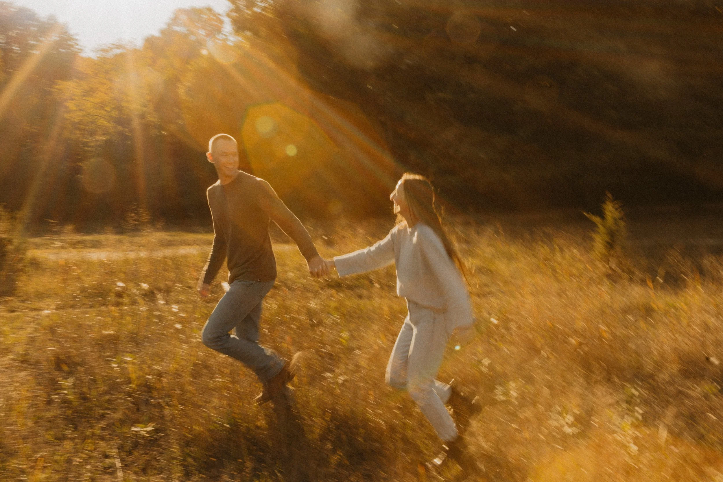A man and woman holding hands and running through a field during sunset, with sunlight creating lens flare effects.