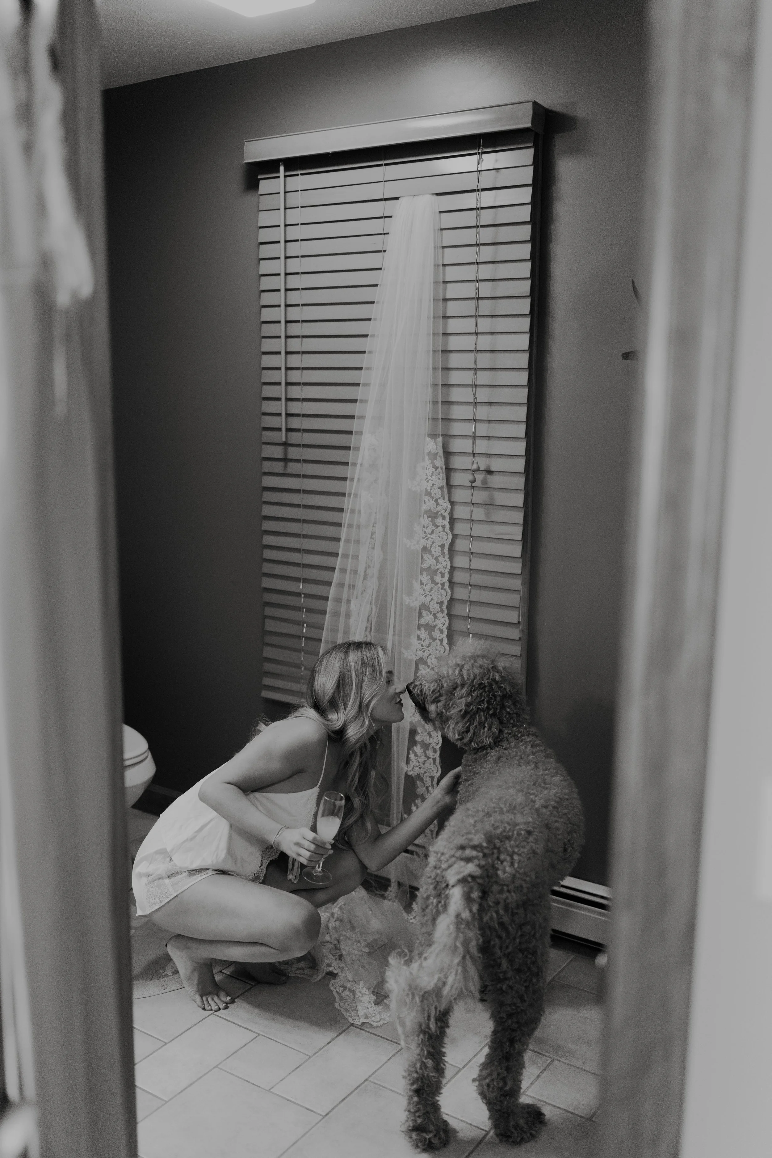 A woman in a white dress holding a glass of champagne kneels and gently kisses a large curly-haired dog in front of a window with blinds and a sheer curtain, as seen through a mirror.