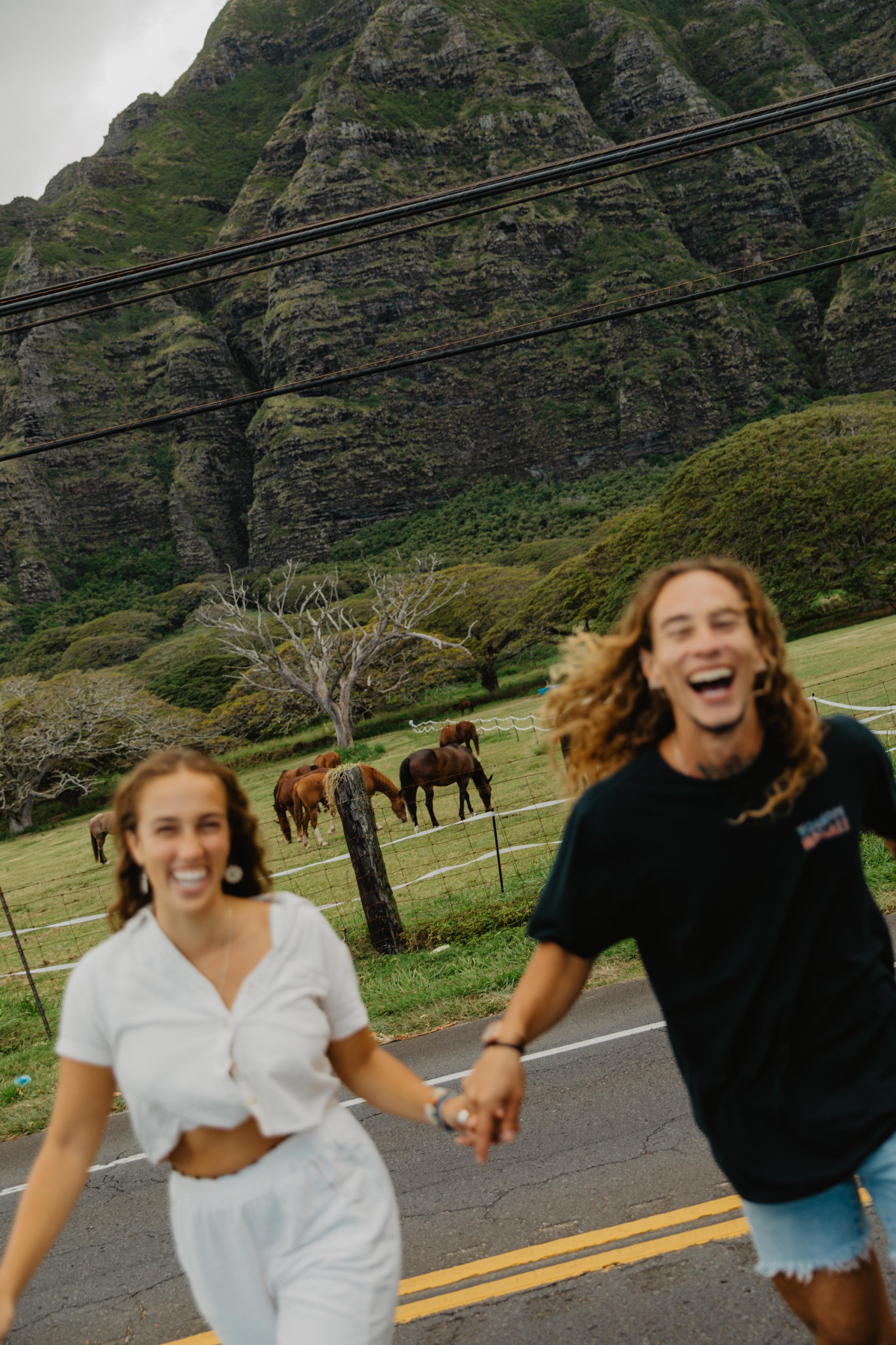 Two young people holding hands and running on a country road with a background of green hills, trees, and horses grazing, with mountains and power lines in the distance.