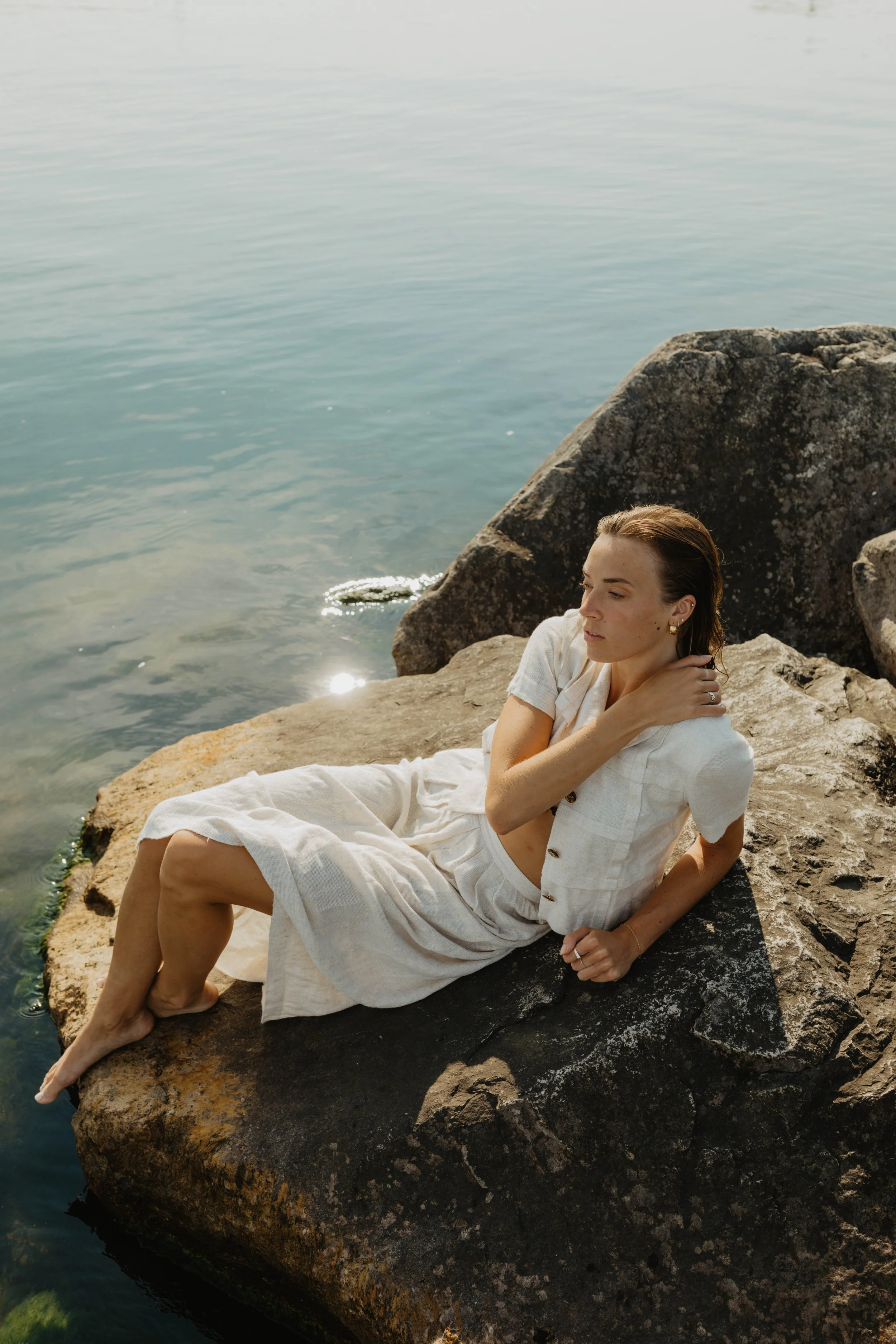 A woman in white linen clothing sitting on a rock by the water.