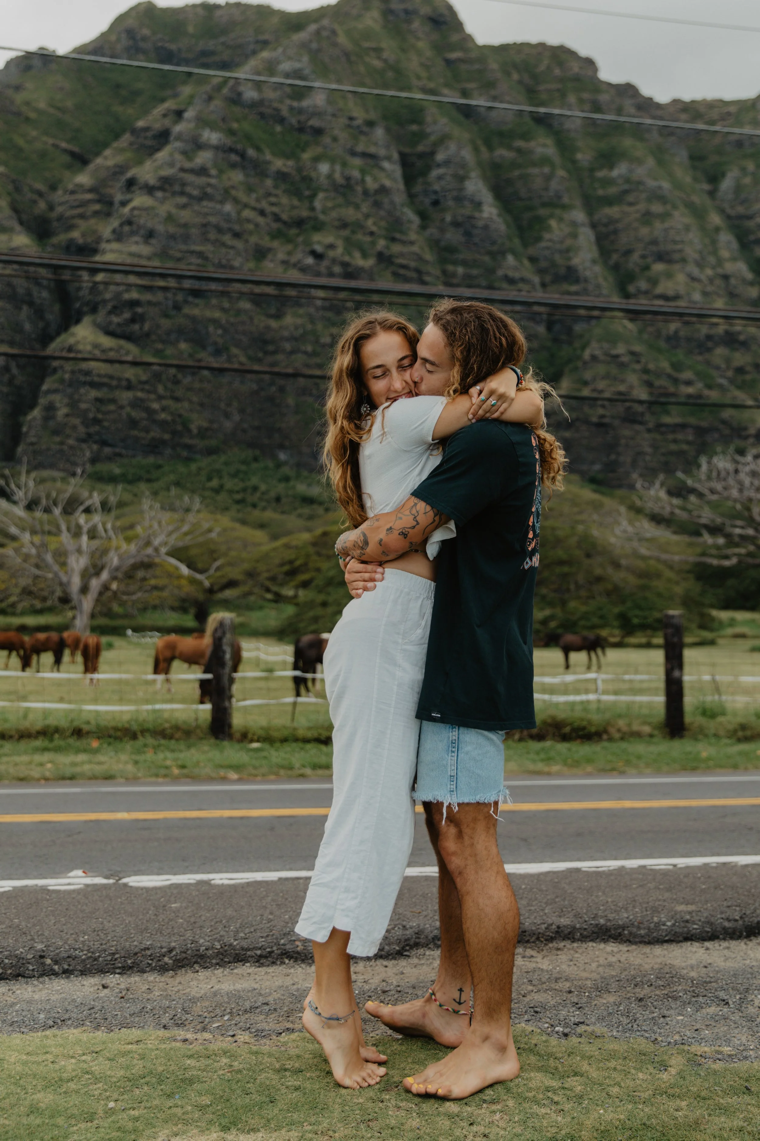 A couple sharing a hug by the side of a rural road with a mountain backdrop and a pasture with horses.
