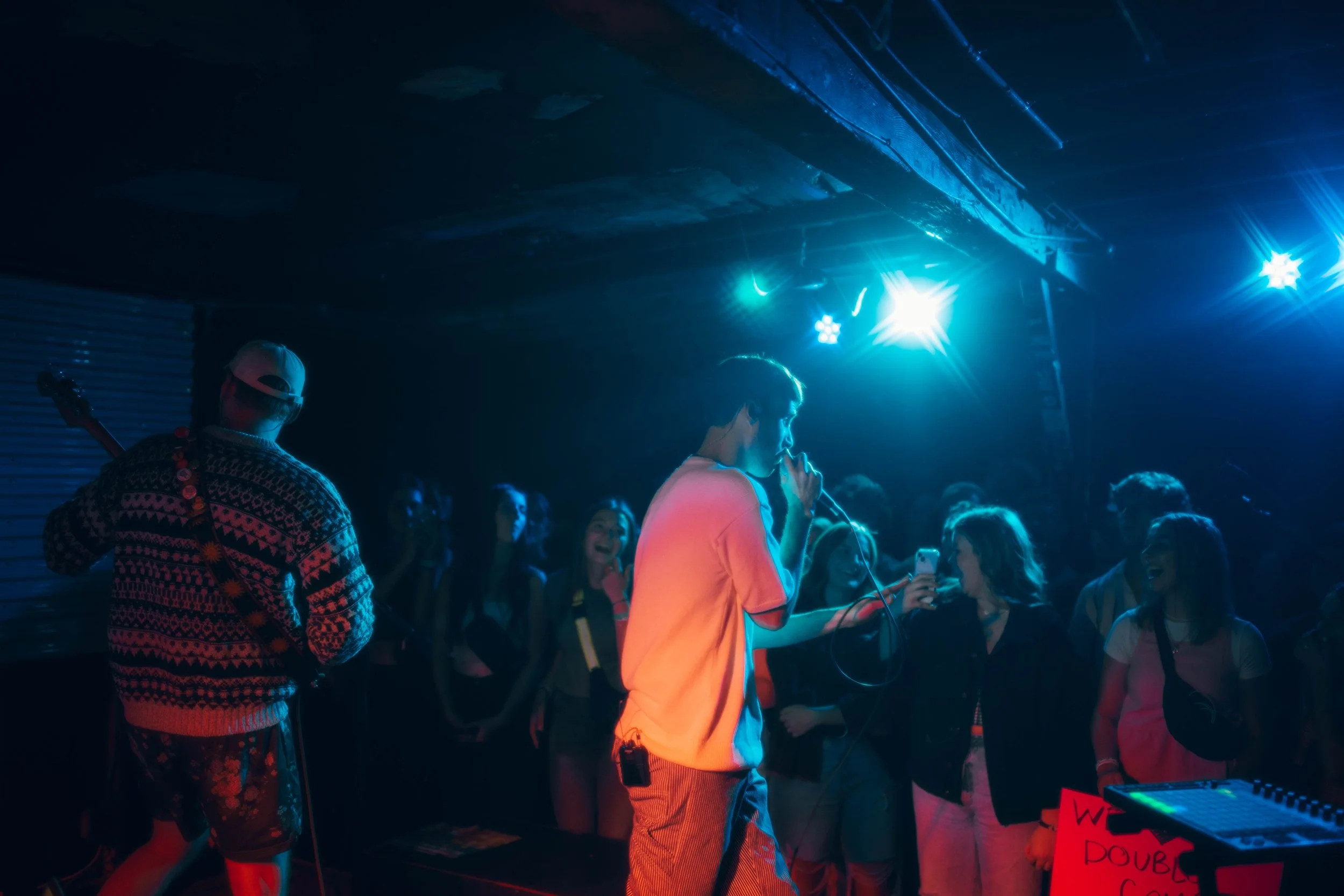 The music artist called DOUBLECAMP  performing with a microphone on stage at a concert, surrounded by a crowd of excited fans. The scene is illuminated with blue and purple stage lights.