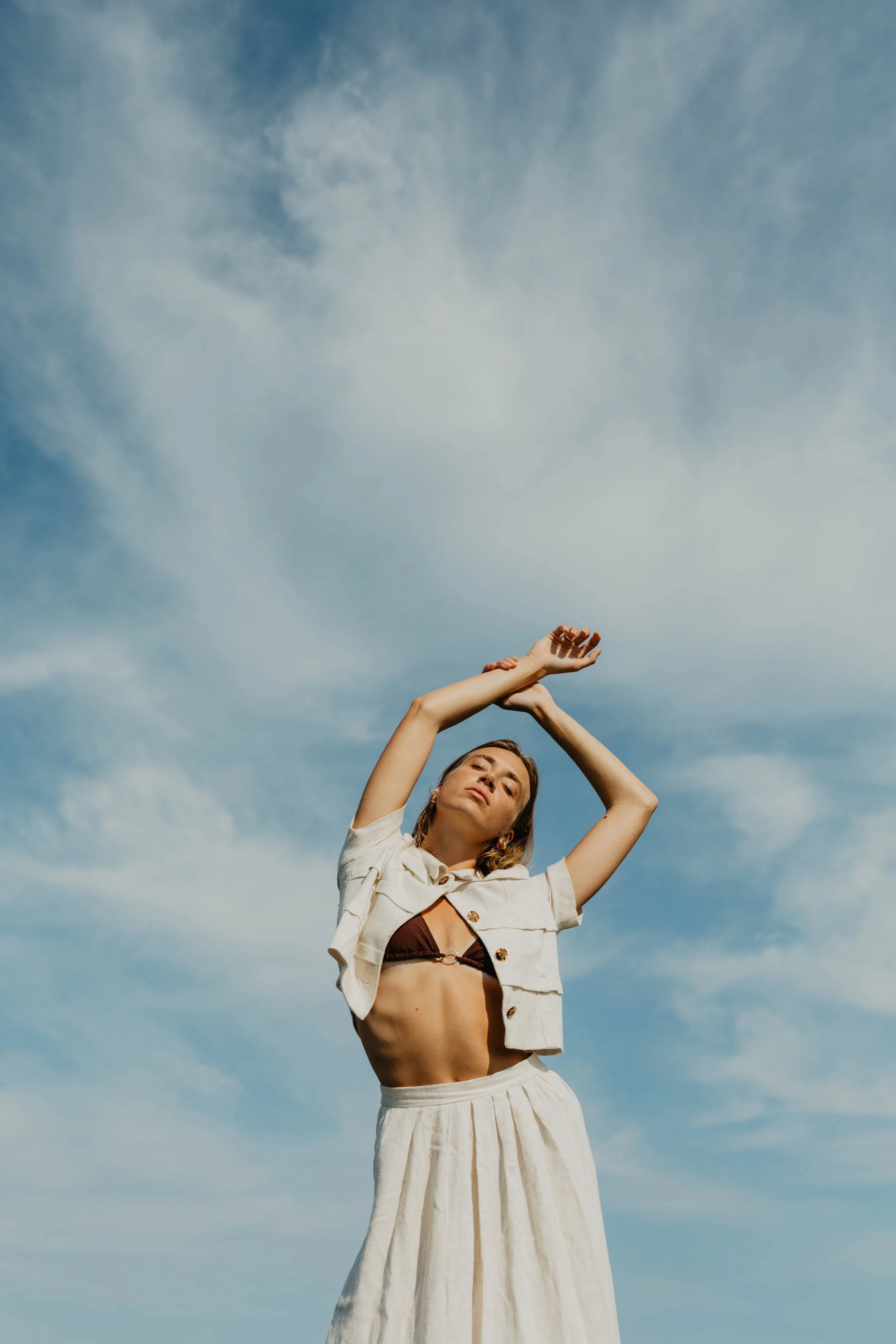 A woman standing outdoors under a blue sky with clouds, wearing a beige two-piece outfit with a brown top, her arms raised above her head, and her eyes closed.