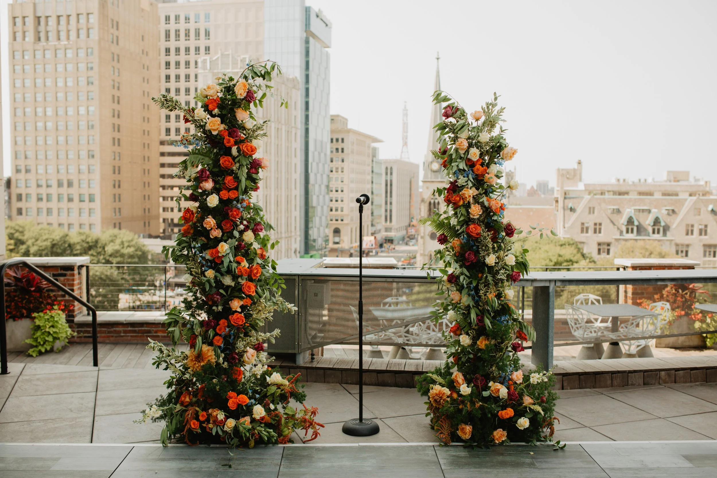 Two tall floral arches with colorful flowers on a balcony overlooking a cityscape with tall buildings and historical architecture.
