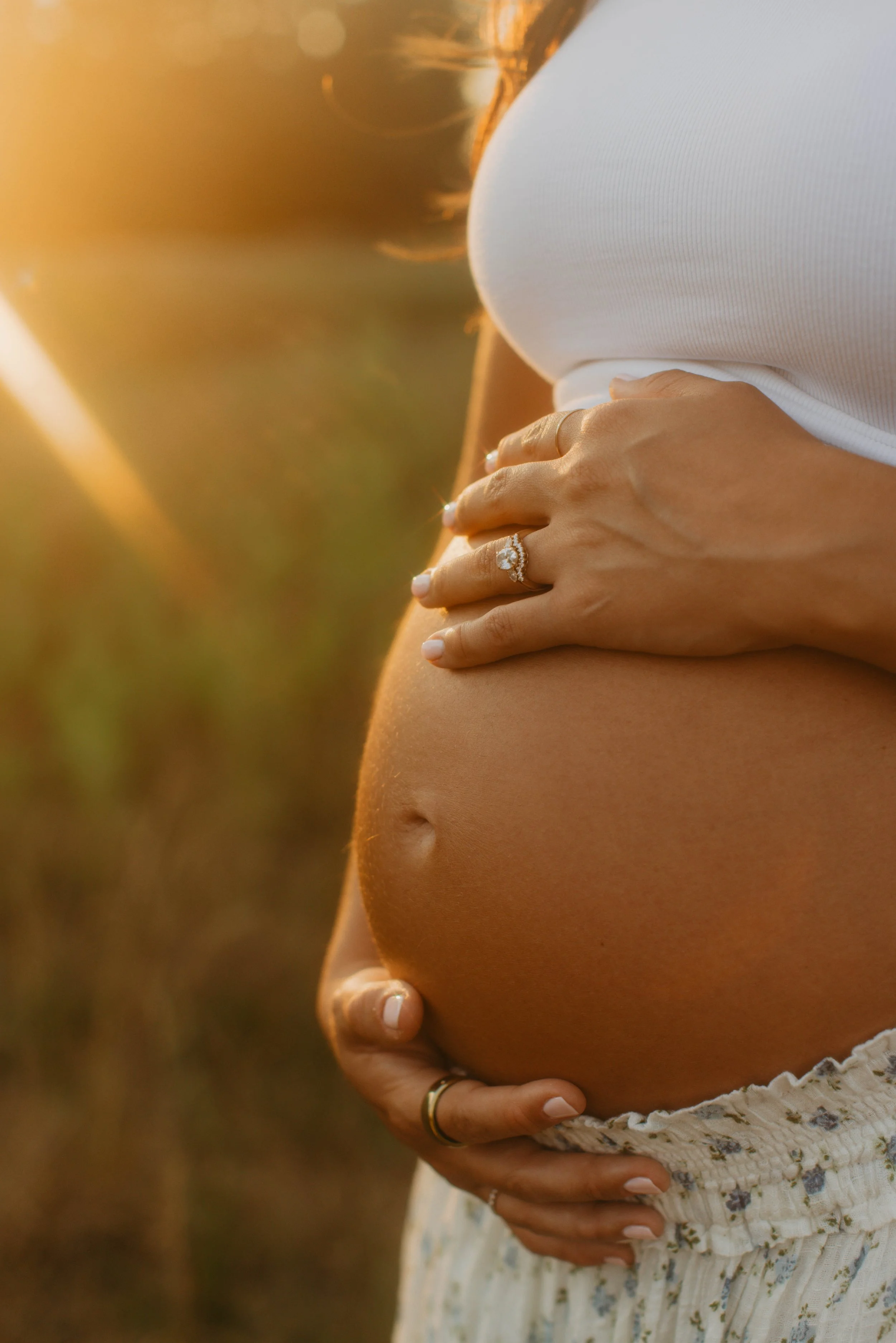 A pregnant woman in a white tank top and patterned skirt is cradling her belly with both hands, one of which has a diamond ring. The photo is taken outdoors during sunset.