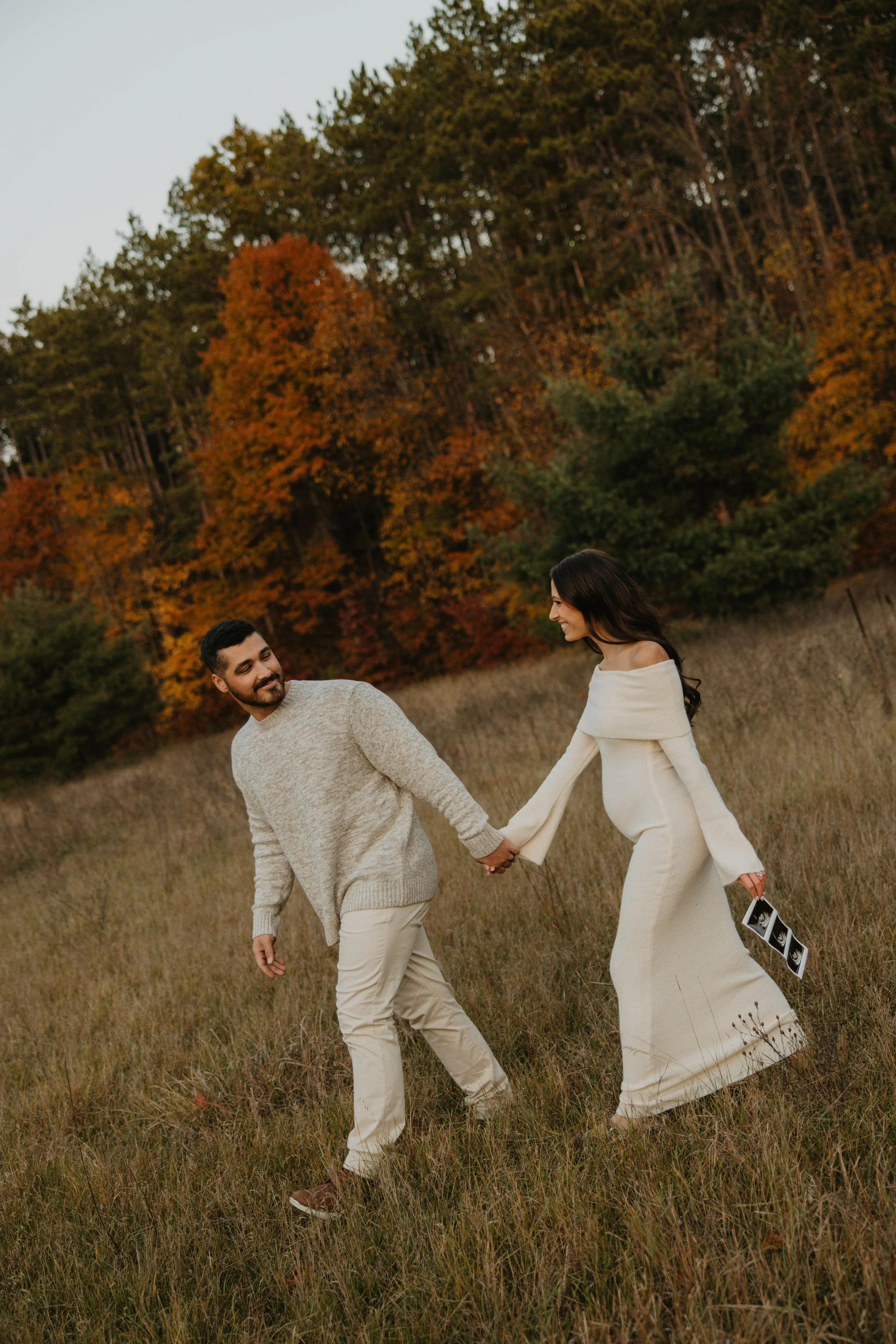 A couple holding hands and walking through a grassy field with autumn trees in the background.