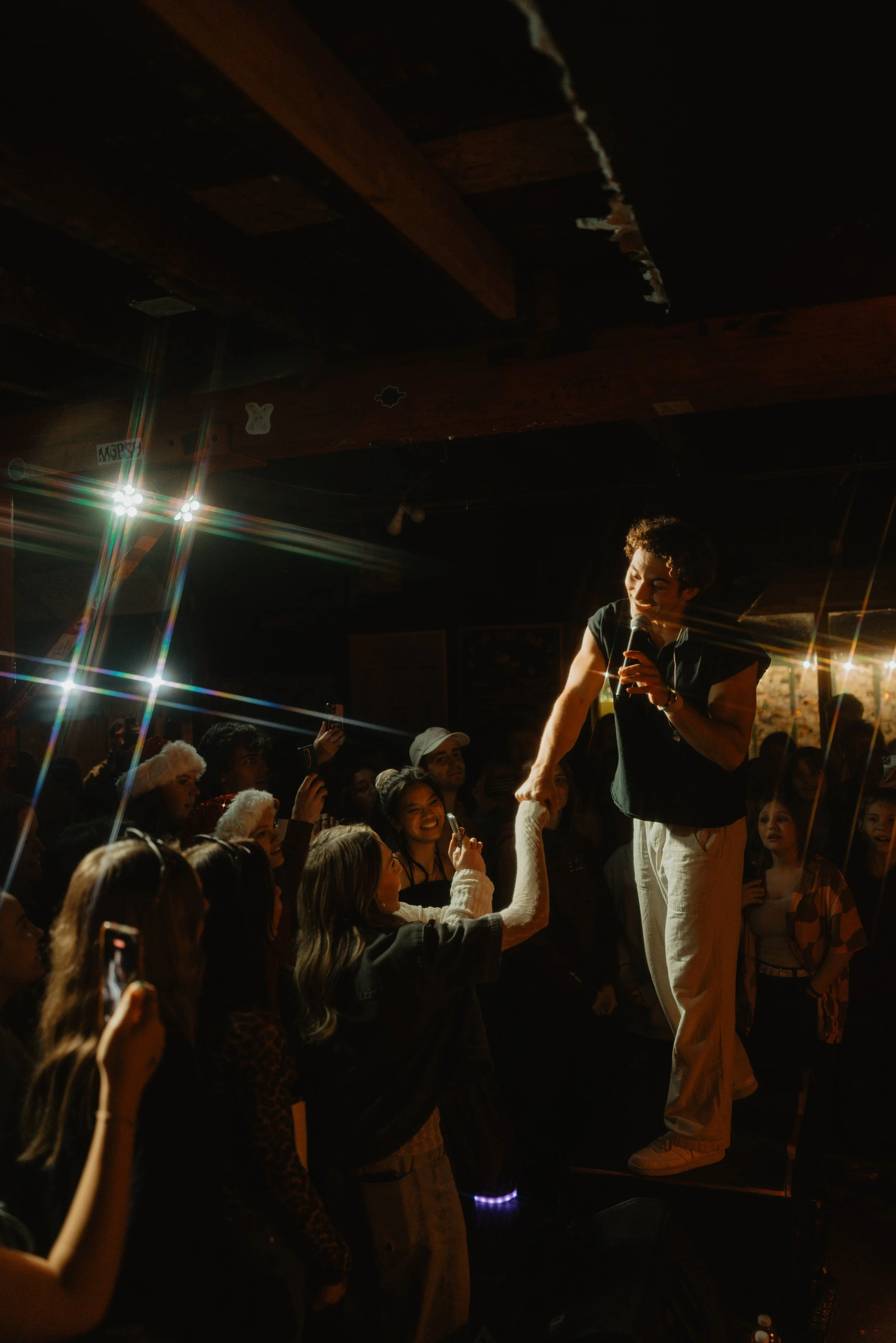 Sammy Rash on stage with a microphone singing to an audience in a dimly lit venue. Audience members enjoy the performance, some taking photos or reaching out for the performer.