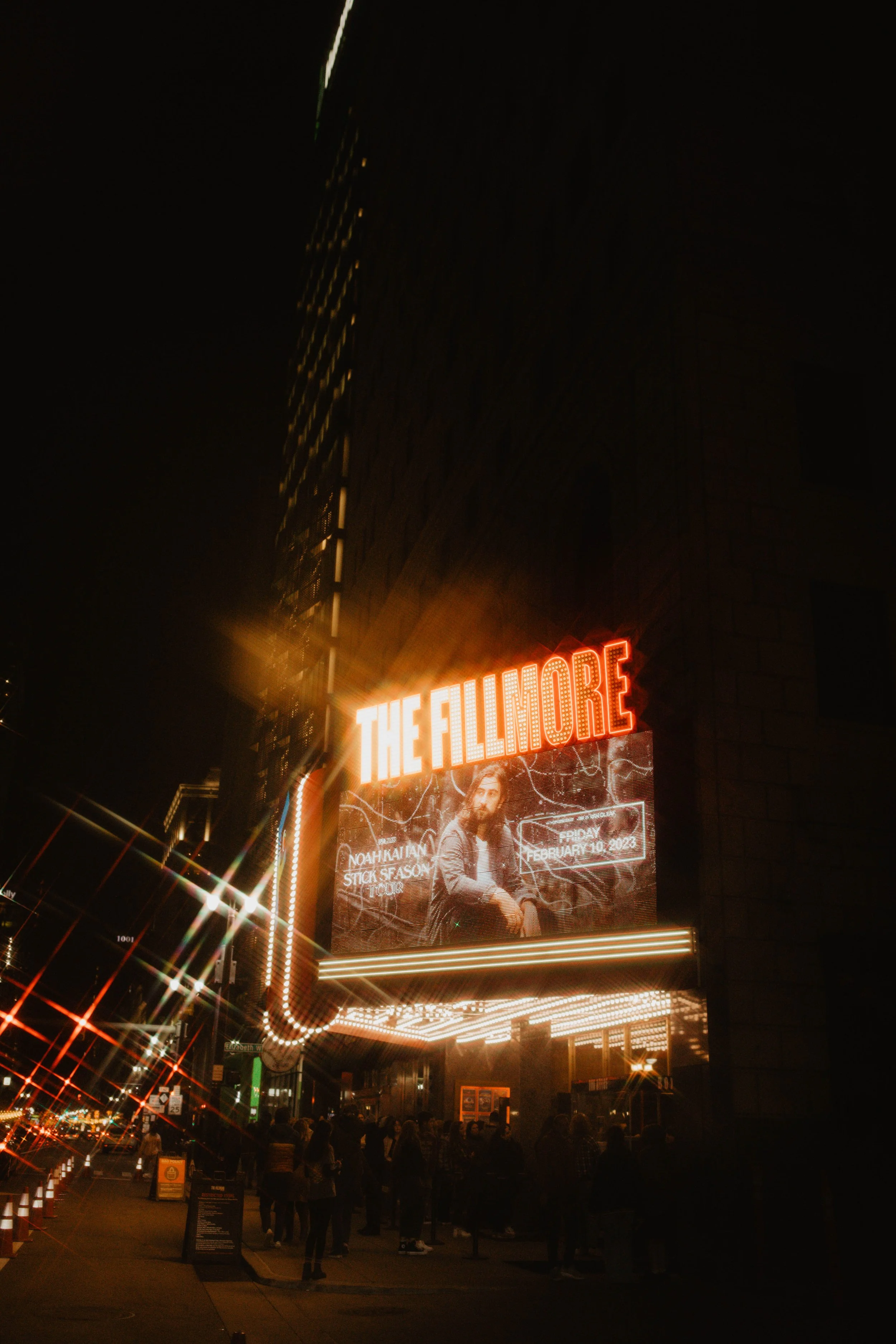 Nighttime view of The Fillmore theater marquee with bright red and white neon lights and an electronic billboard displaying a concert advertisement for Noah Kahan's Stick Season Tour on February 10, 2023, with people gathered outside.