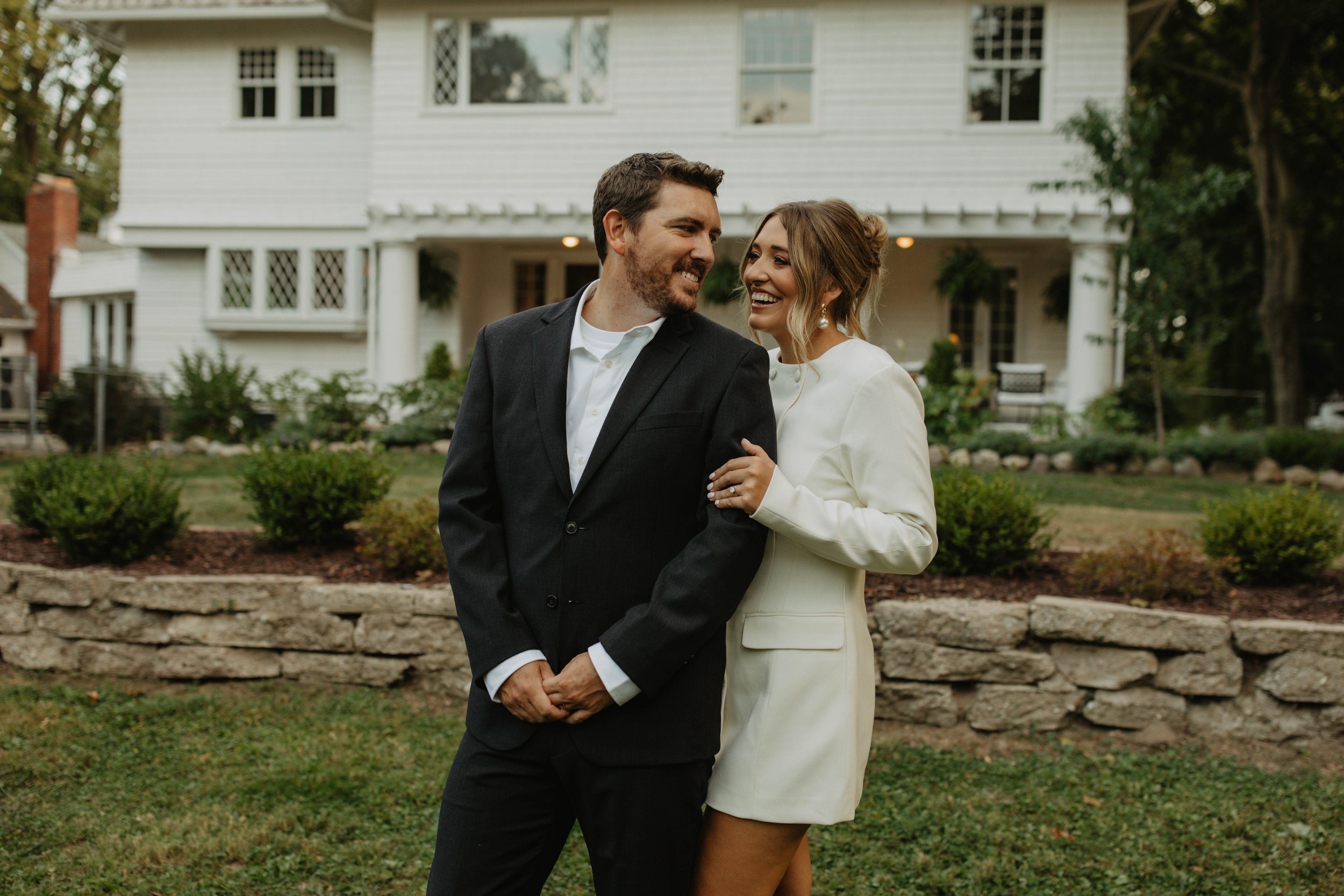 A smiling couple stands together on a lawn in front of a large white house, with the woman wearing a white dress and the man in a black suit, sharing a loving moment.