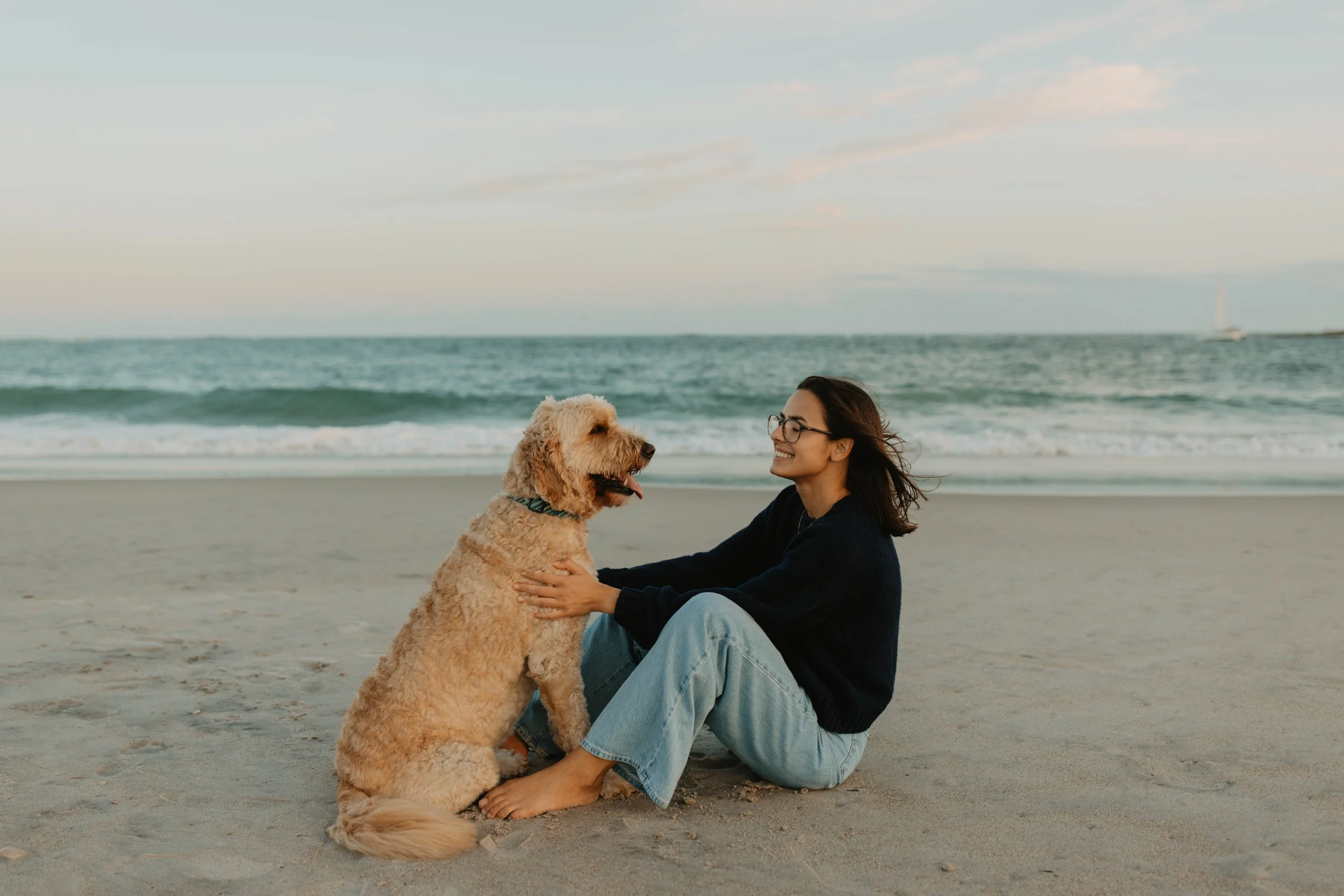 A woman sitting on the sand at the beach, smiling and holding a large, curly-haired dog that is sitting beside her, with ocean waves in the background.