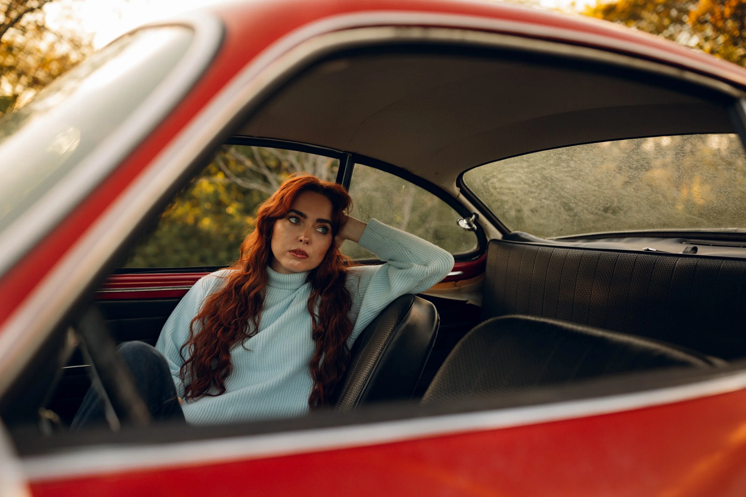A woman with long red hair sitting inside a vintage red car, looking out the window with a contemplative expression, during autumn with fall foliage outside.