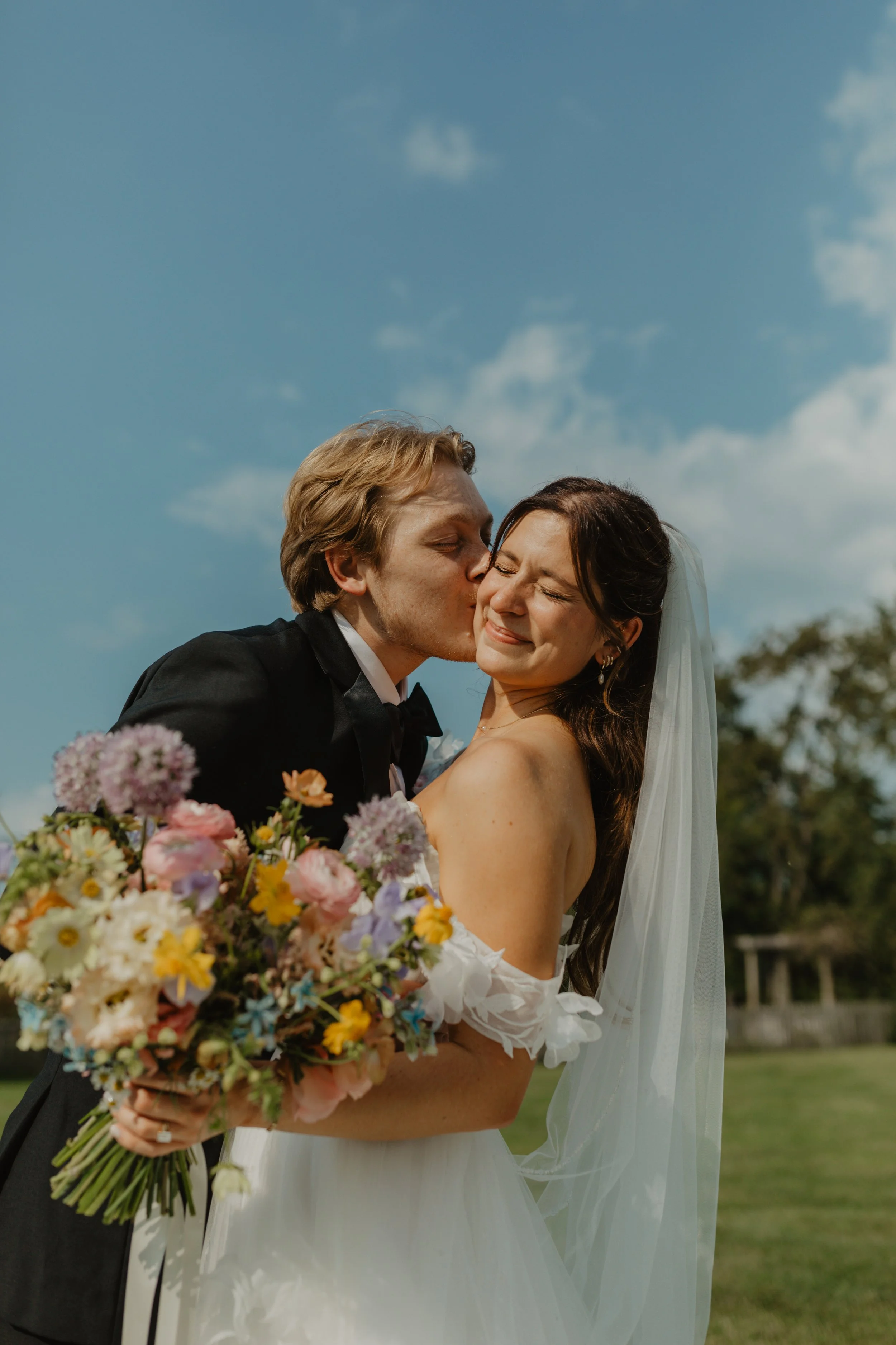 A newlywed couple on their wedding day, with the groom kissing the bride on her cheek while she smiles with her eyes closed, holding a bouquet of colorful flowers outdoors under a blue sky.
