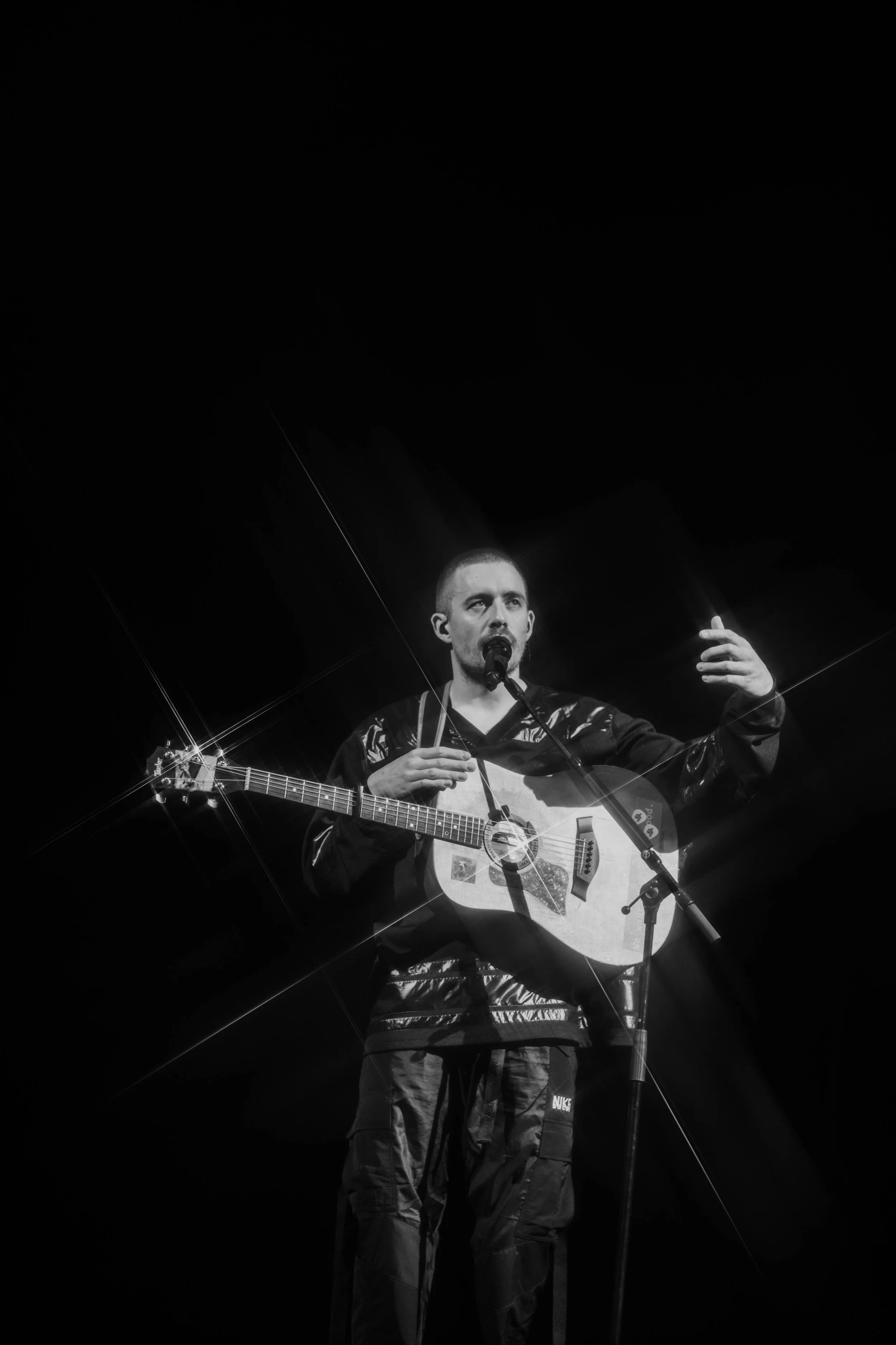 A man performing on stage plays an acoustic guitar and sings into a microphone, gesturing with his right hand, against a dark background.