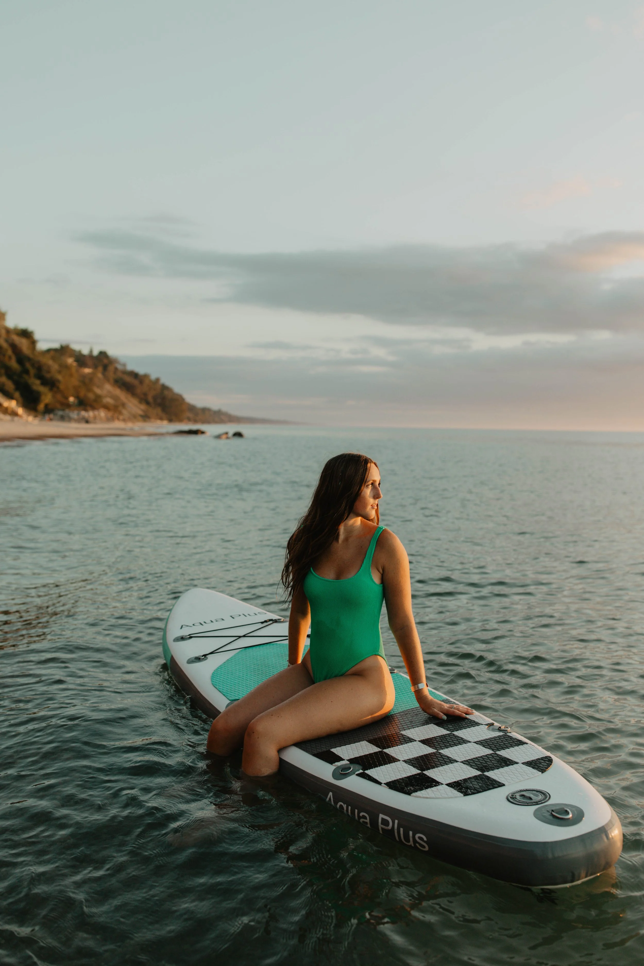 A woman in a green swimsuit sitting on a paddleboard in the water during sunset, with a shoreline and cloudy sky in the background.