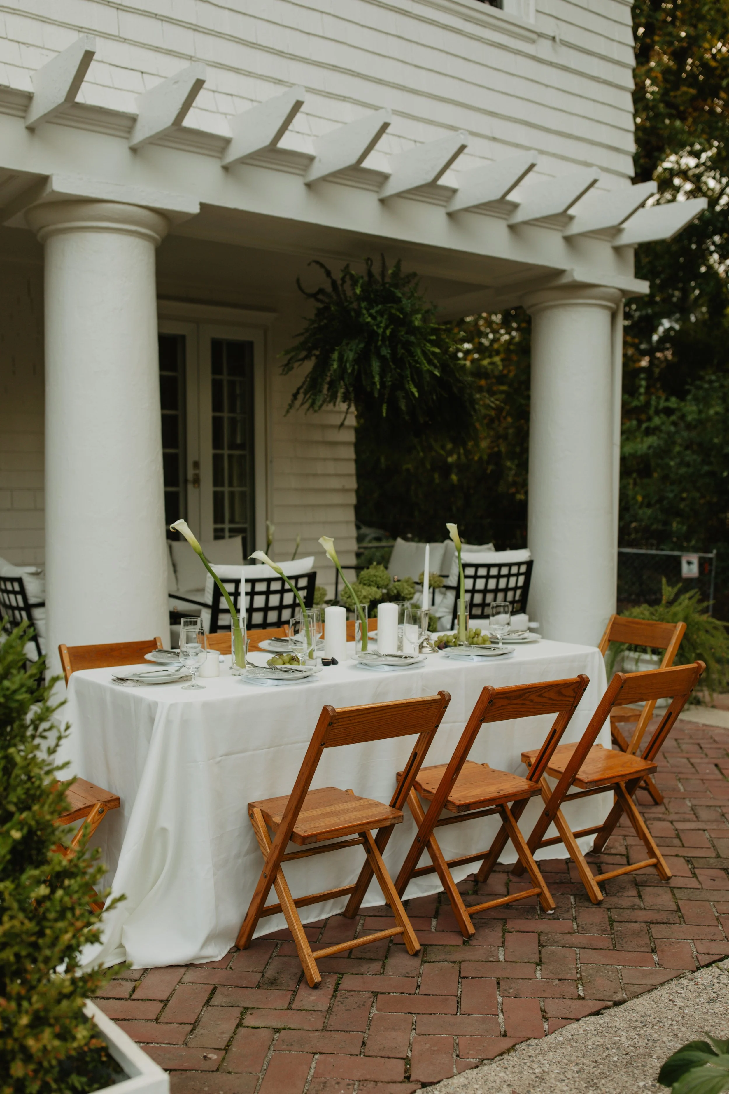 An outdoor patio with a dining table set for a meal, featuring a white tablecloth, candles, flowers, and glassware, surrounded by wooden chairs, under a porch with white columns and a hanging plant.