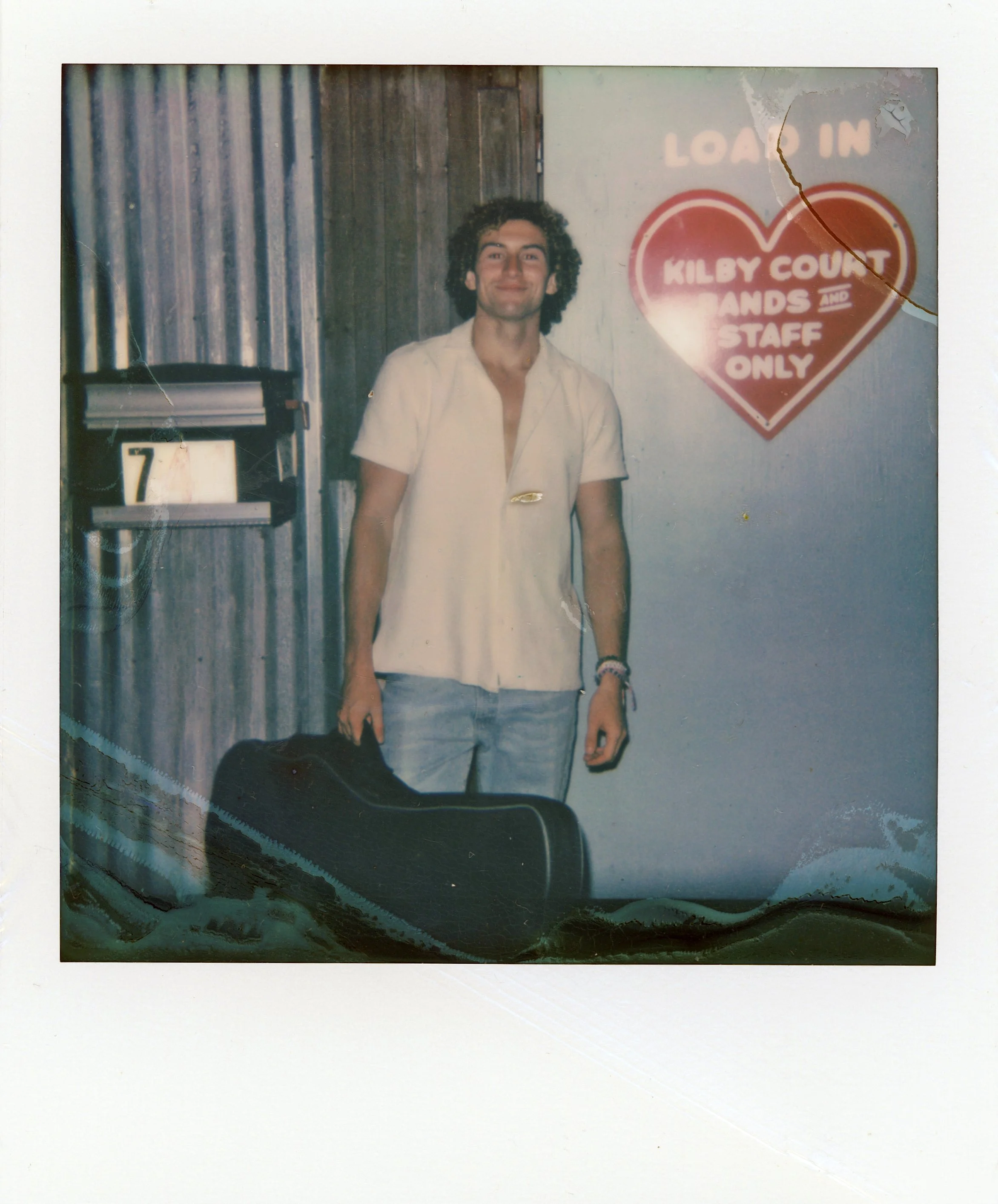 Sammy Rash, standing indoors near a wooden wall and a sign that says 'LOAD IN' and 'KILBY COURT HANDS AND STAFF ONLY' in a heart-shaped design.