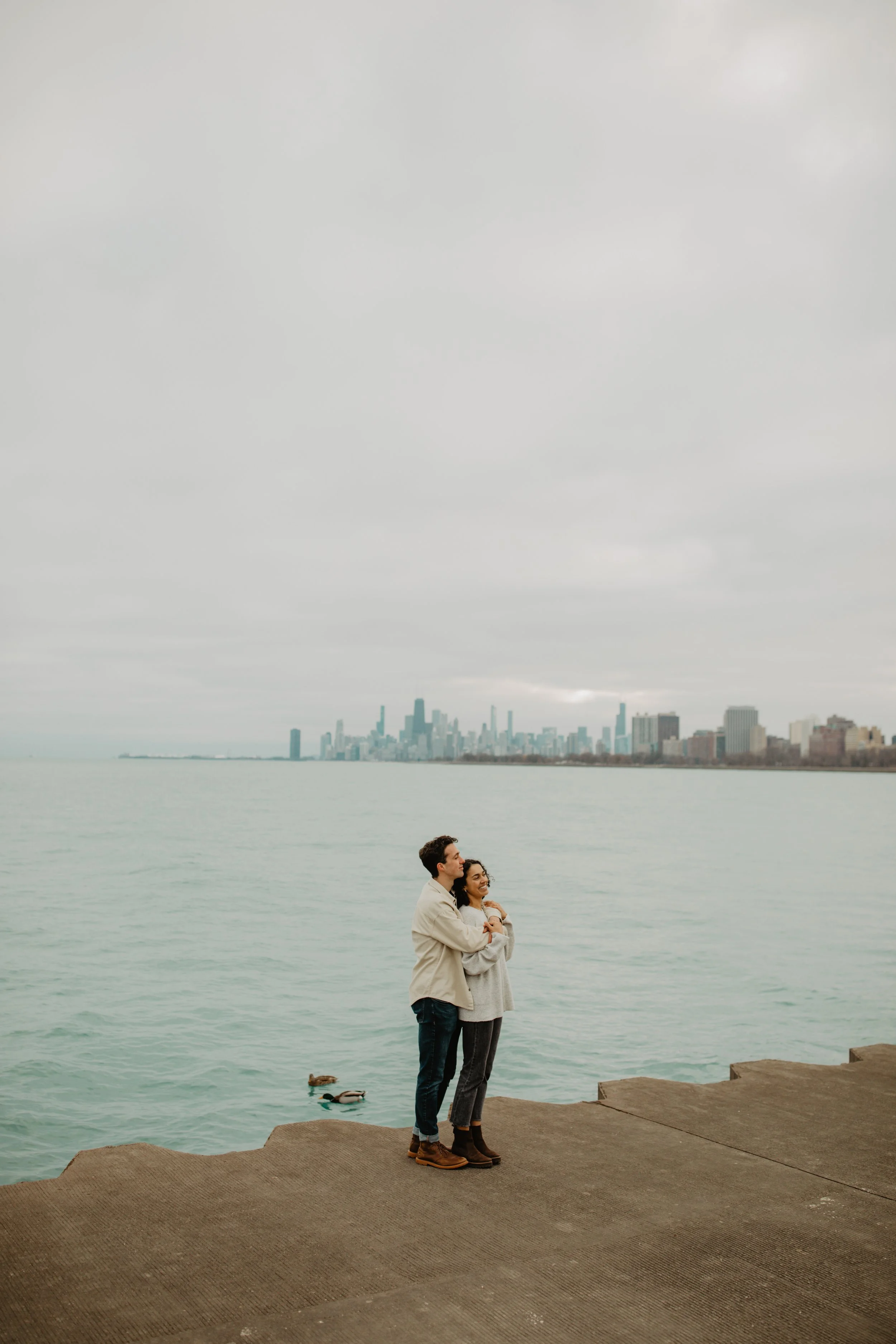 A couple standing on a concrete pier hugging each other with a city skyline in the background and ducks in the water nearby.