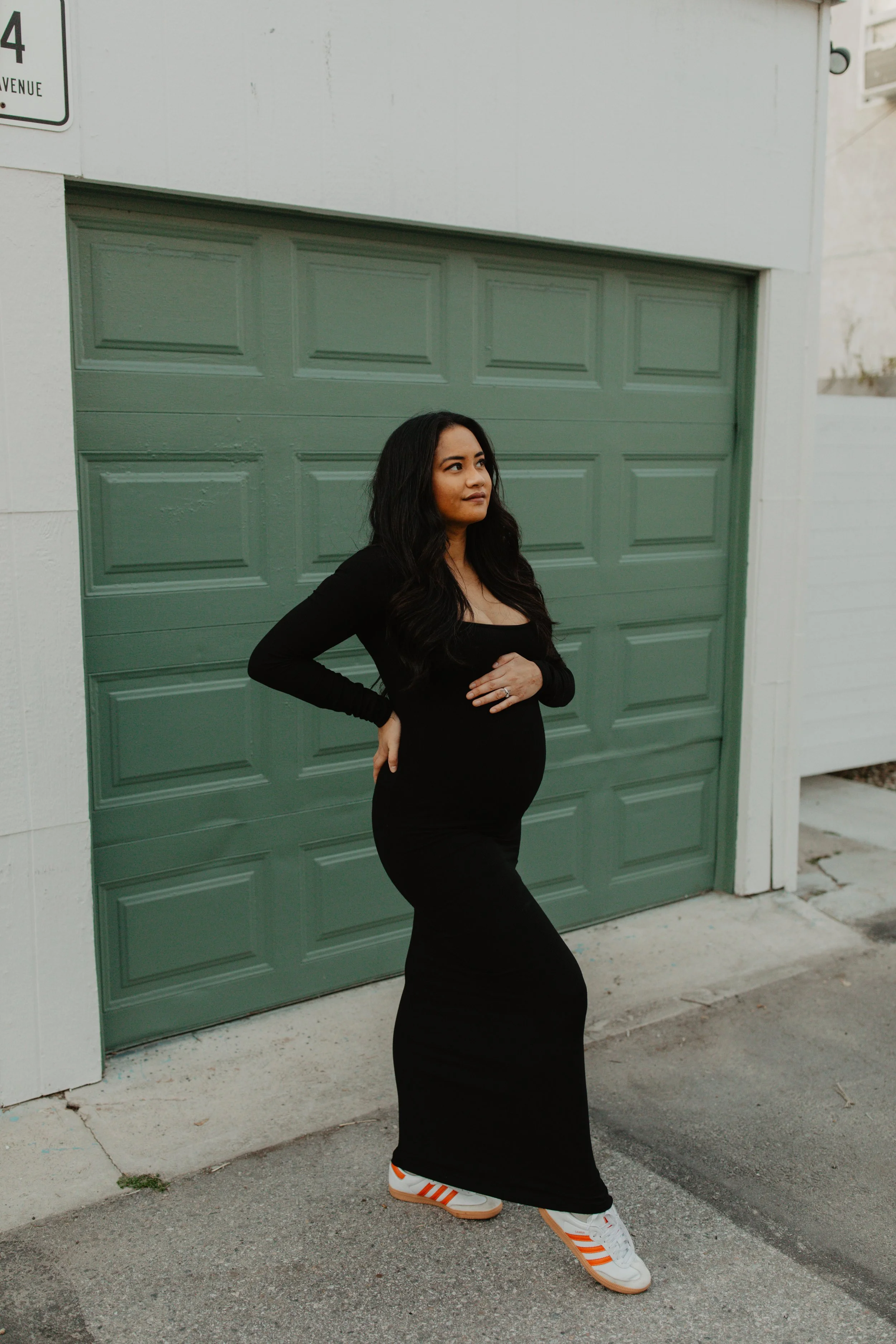 Pregnant woman in a black dress and sneakers standing in front of a green garage door on a street.