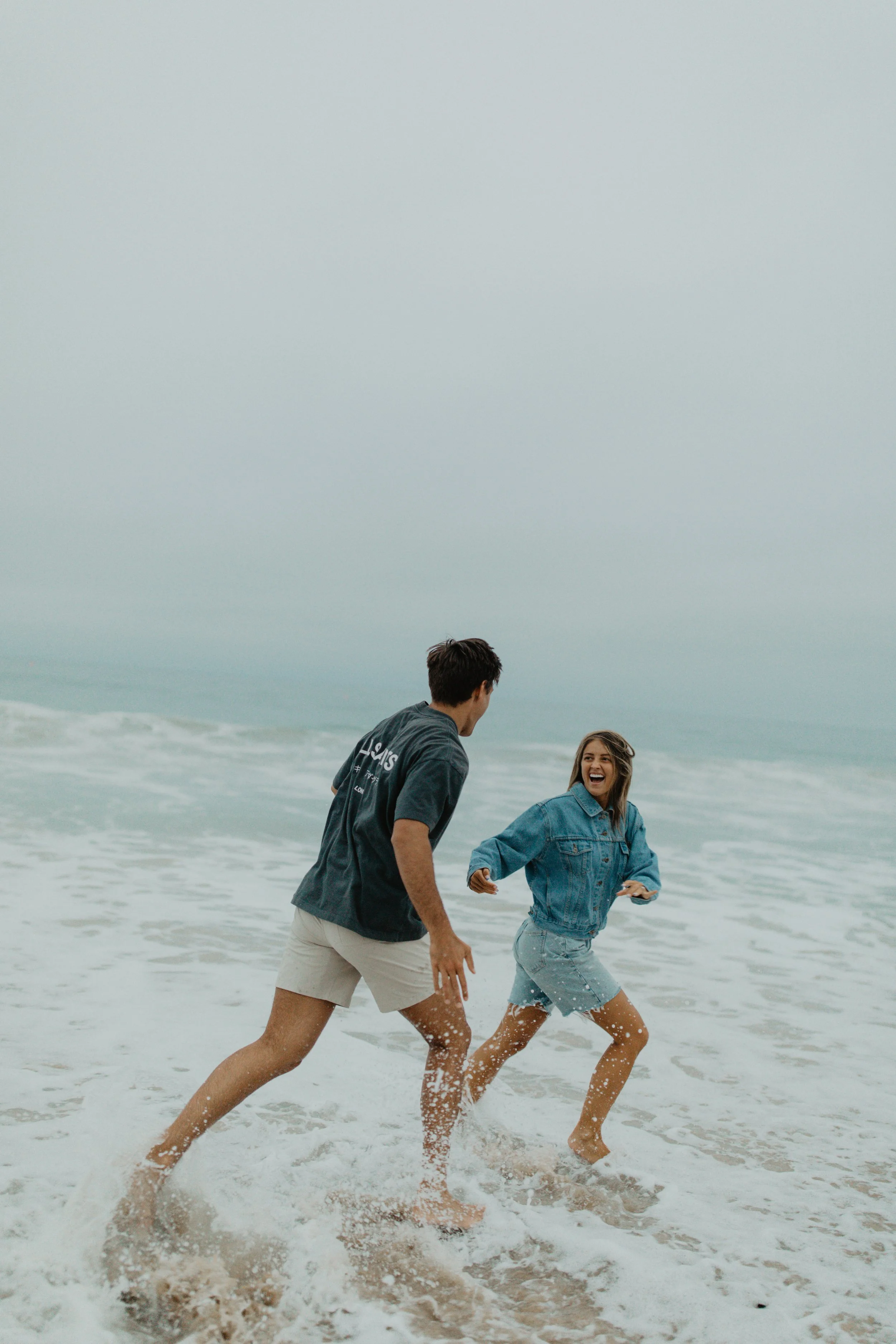 A couple playfully running in the ocean waves at the beach on a foggy day, laughing and enjoying themselves in California.