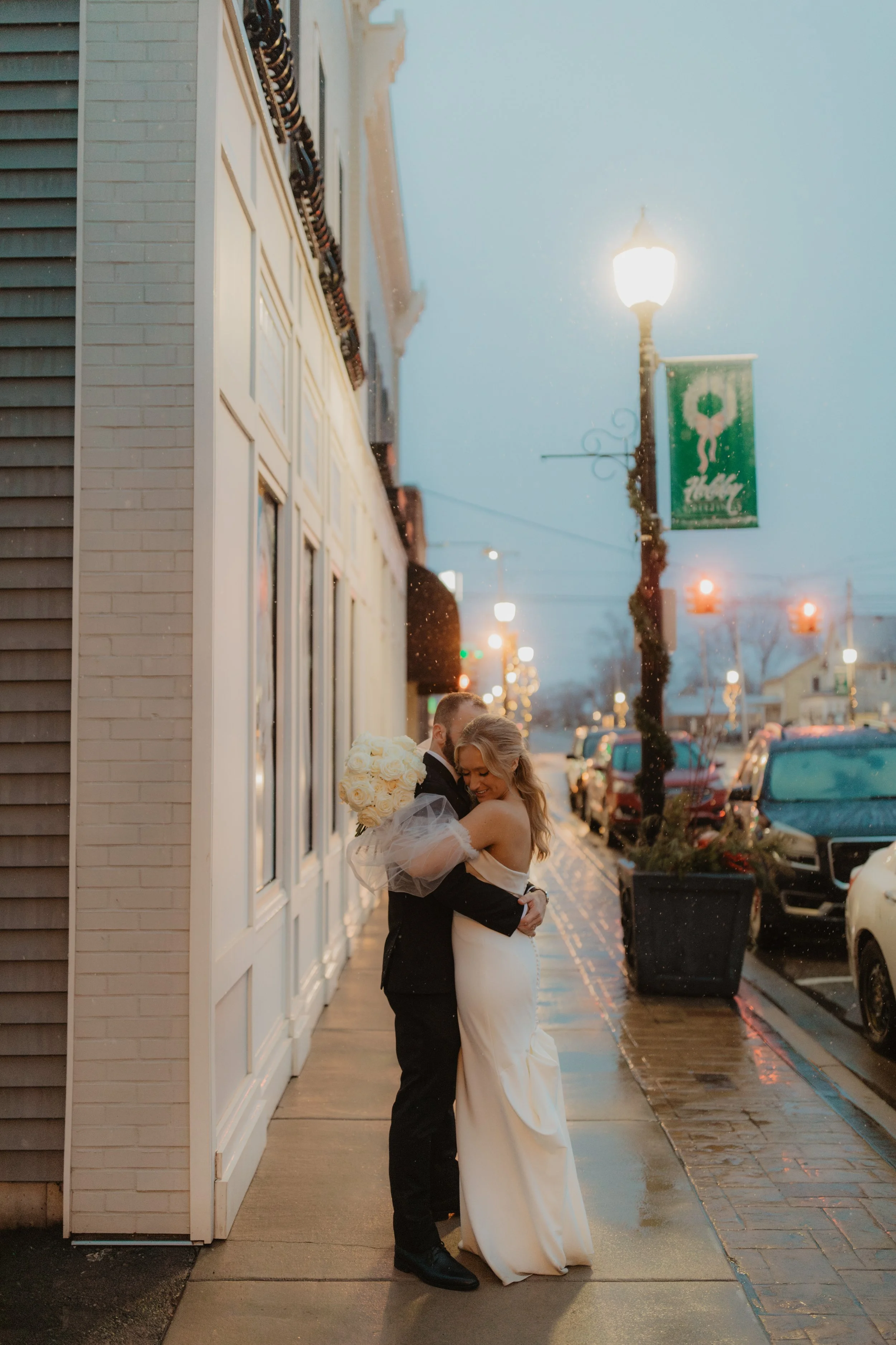 A bride and groom hugging on a rainy city sidewalk at dusk, with cars parked nearby and decorative streetlights.
