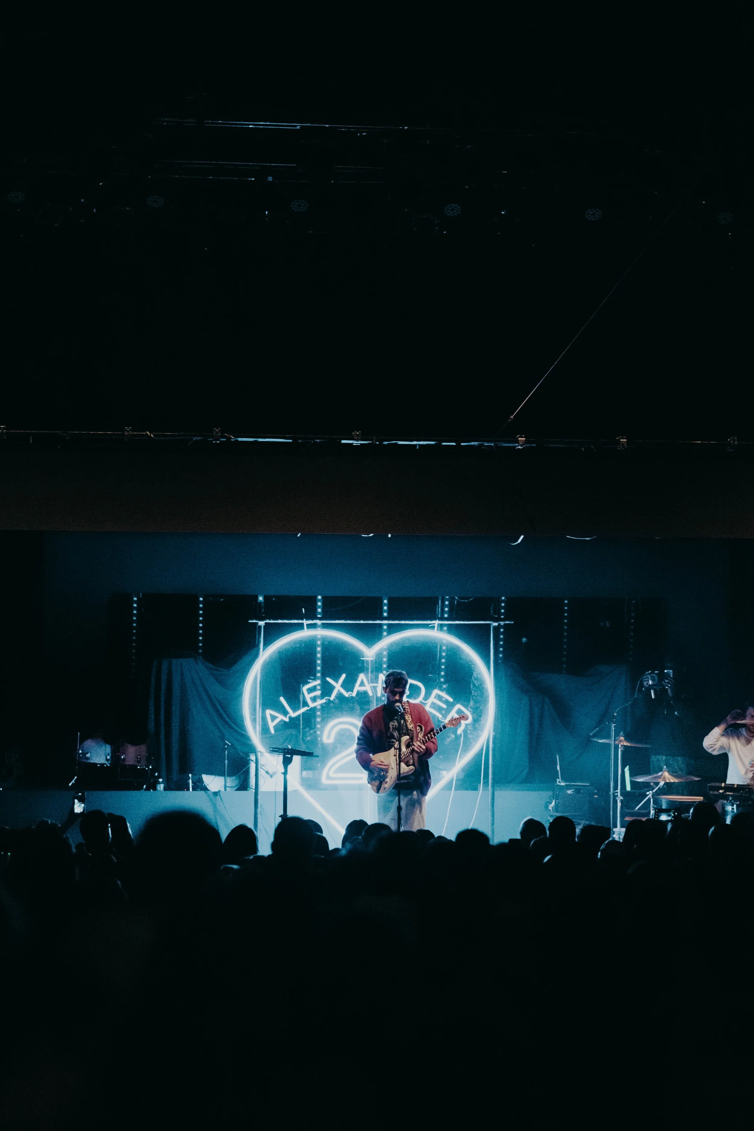 Alexander22 on stage with a neon sign behind the performer reading 'ALEXANDER' in a heart shape, with band members playing instruments.