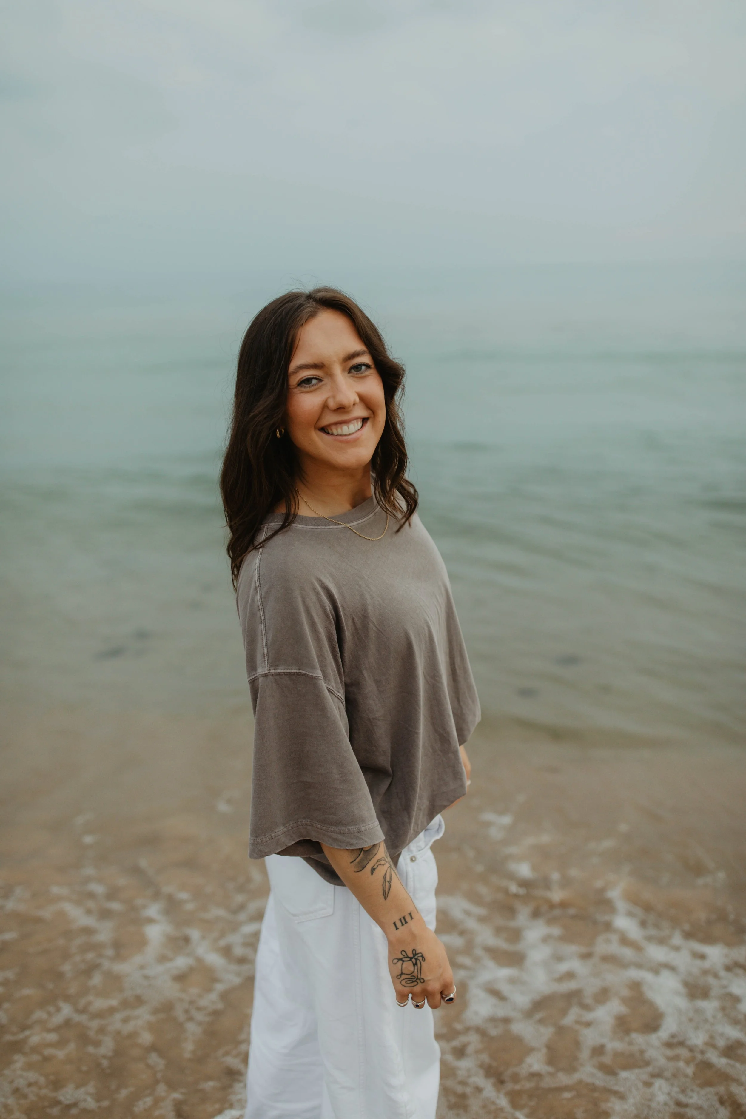 A woman standing on a beach with a smile, wearing a gray t-shirt and white pants, with tattoos on her arm, in front of the ocean under a cloudy sky.