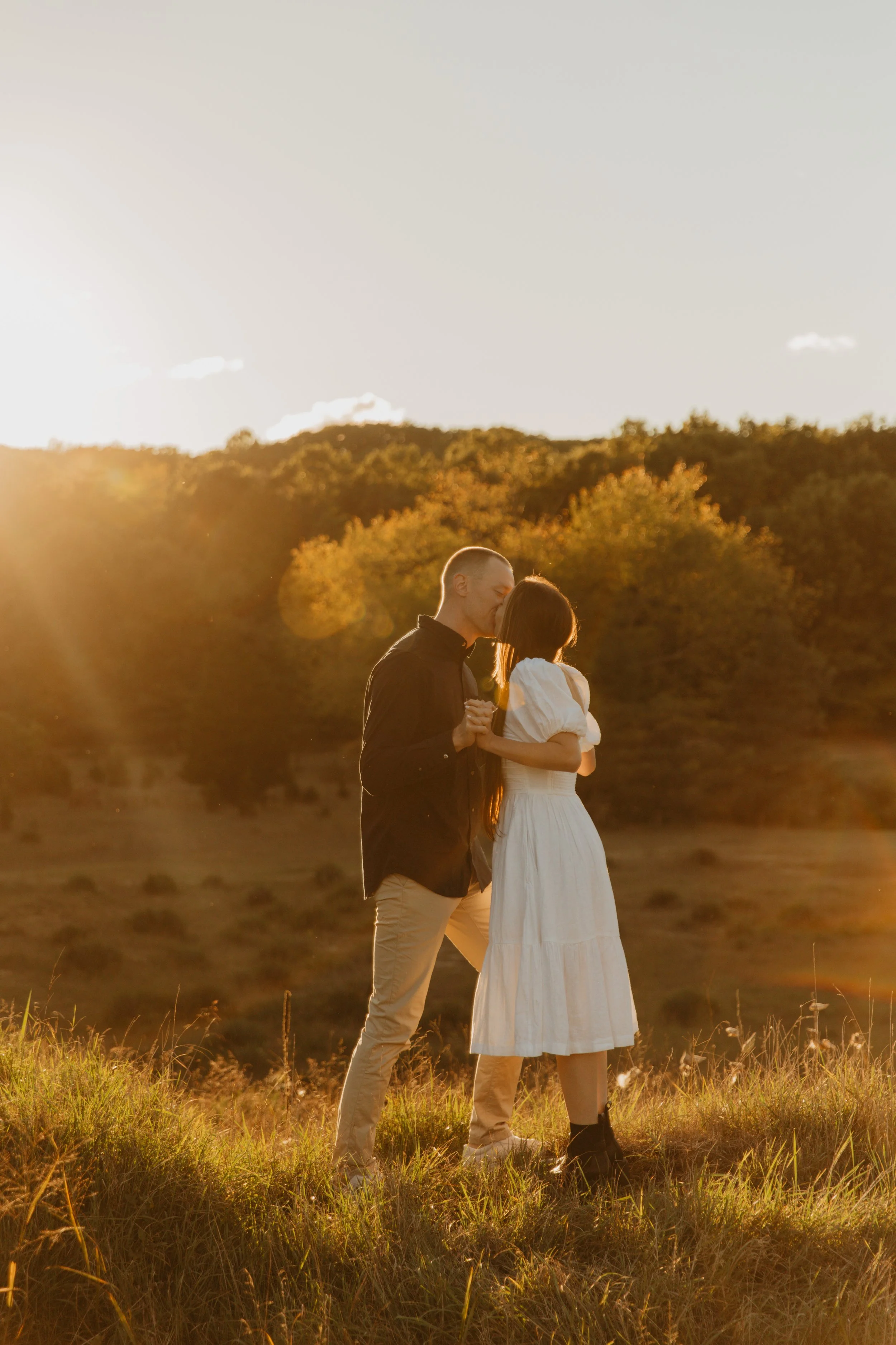 A couple kissing outdoors during golden hour, with autumn trees in the background and sunlight creating a warm glow.