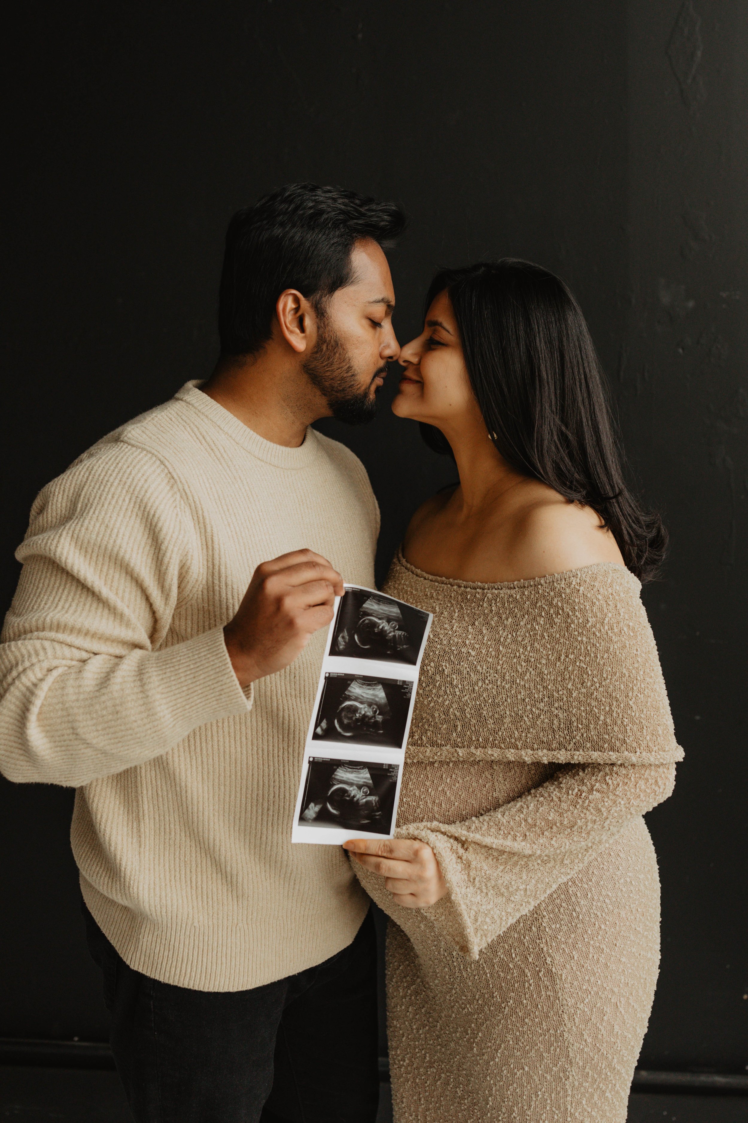 A couple holding ultrasound images, standing close together, about to kiss, against a black background.