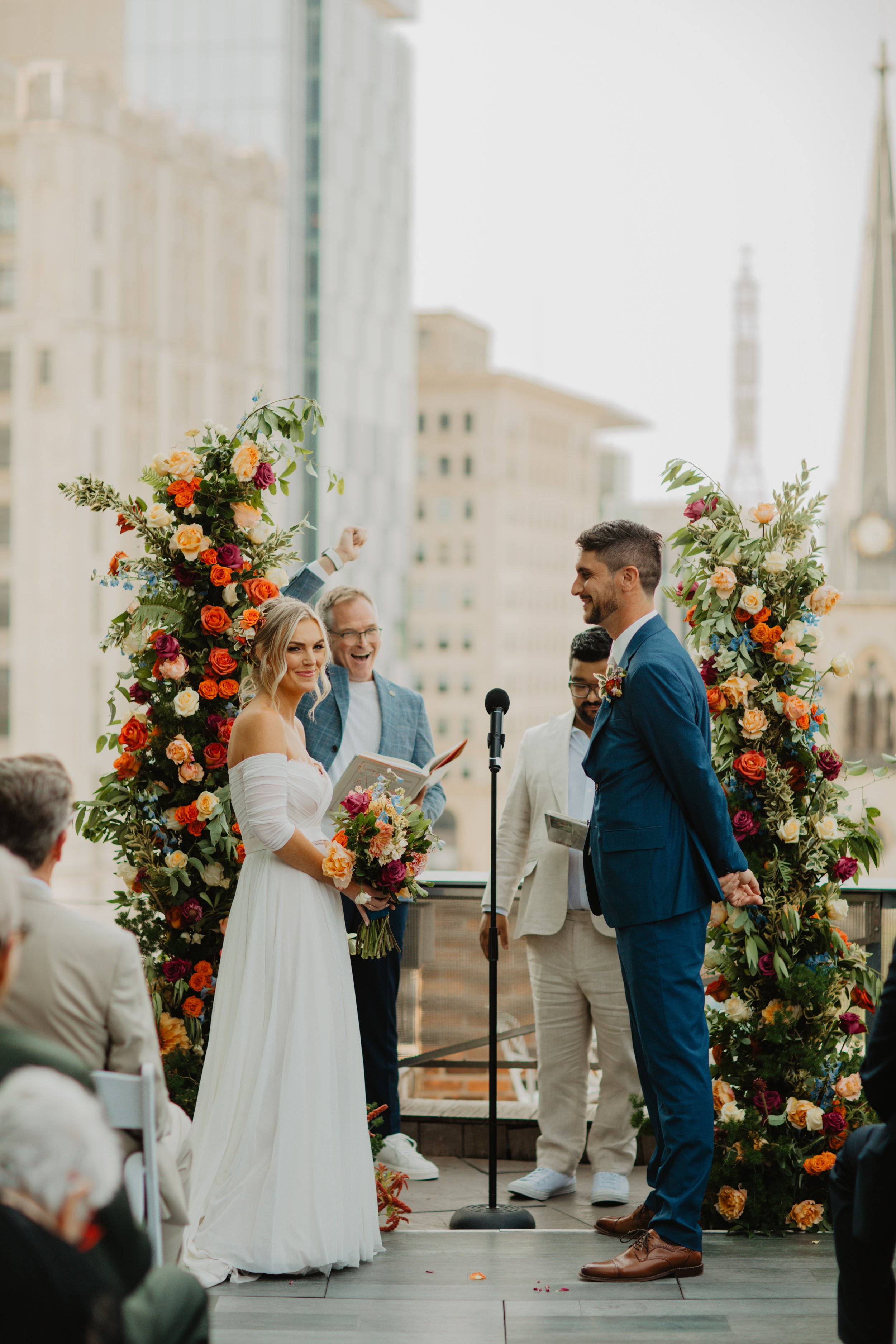 A bride and groom stand facing each other during their wedding ceremony on a rooftop, surrounded by floral arrangements. The bride holds a bouquet and reads vows, while the groom listens smiling. Officiant in the background reads from a book, with gu