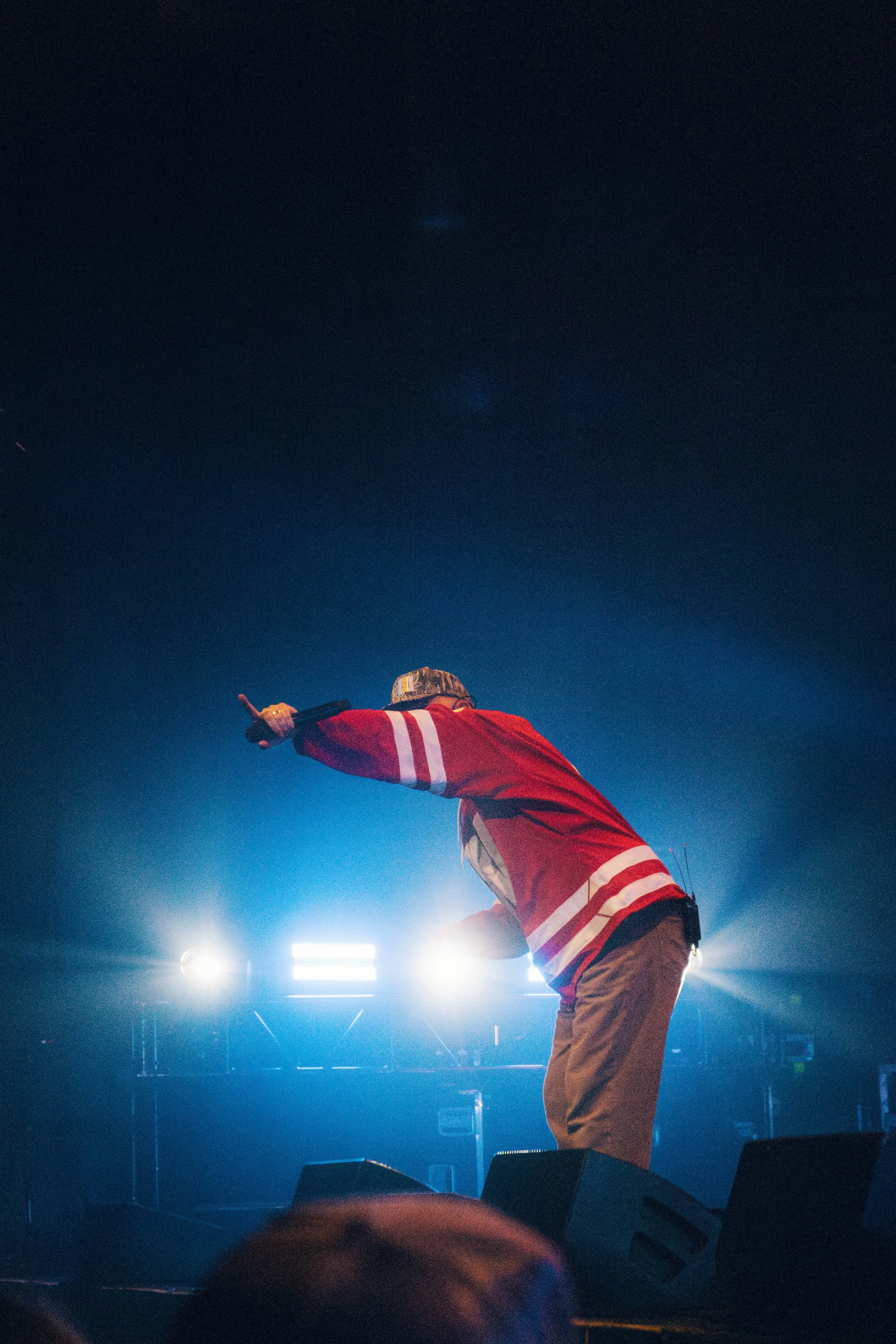 The music artist Quinn XCII performing on stage, wearing a red jacket with white stripes, bending forward and pointing with one hand, with bright stage lights behind them.