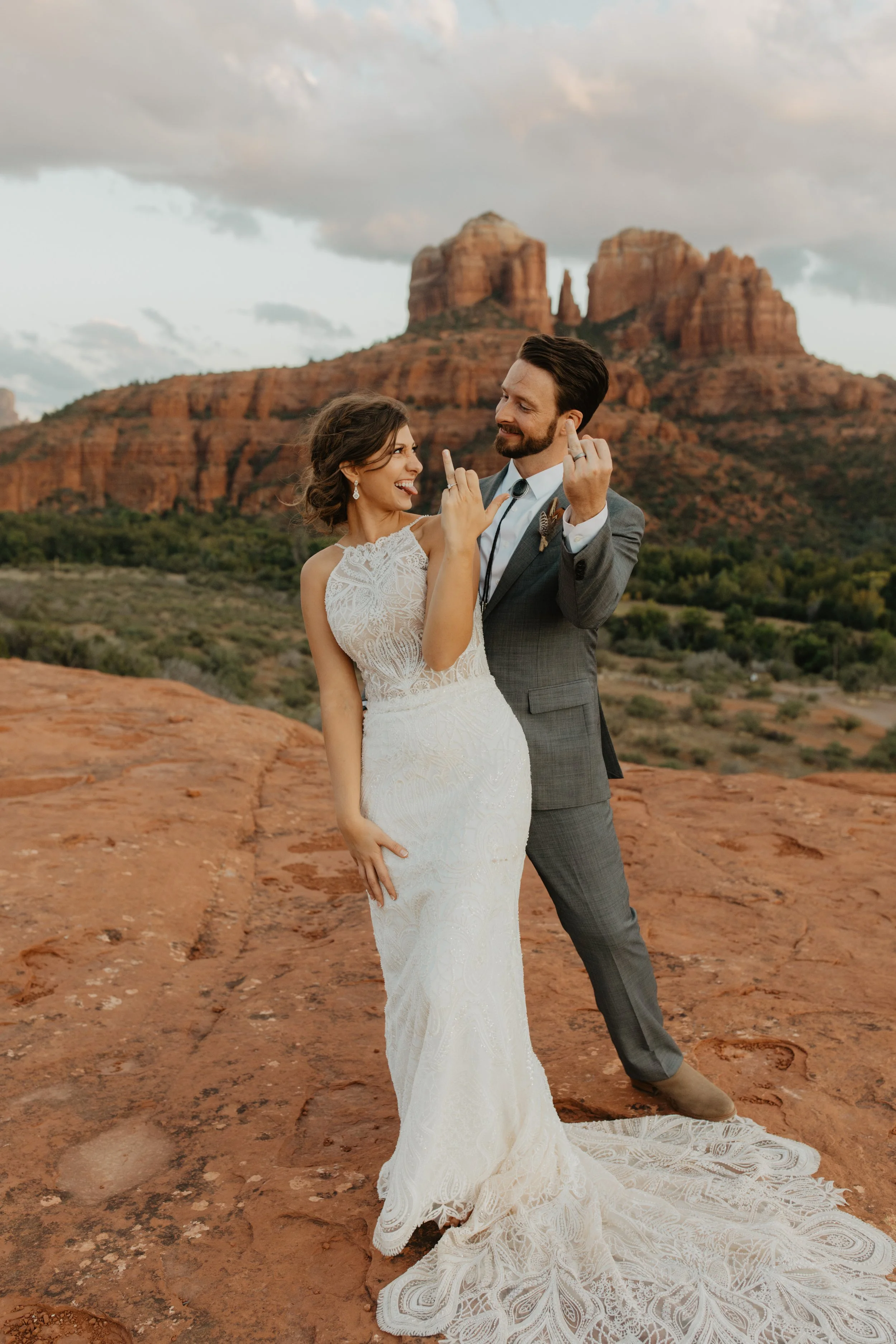 A couple in wedding attire standing on a red rock landscape at sunset, with a canyon and rock formations in the background, smiling and touching hands.