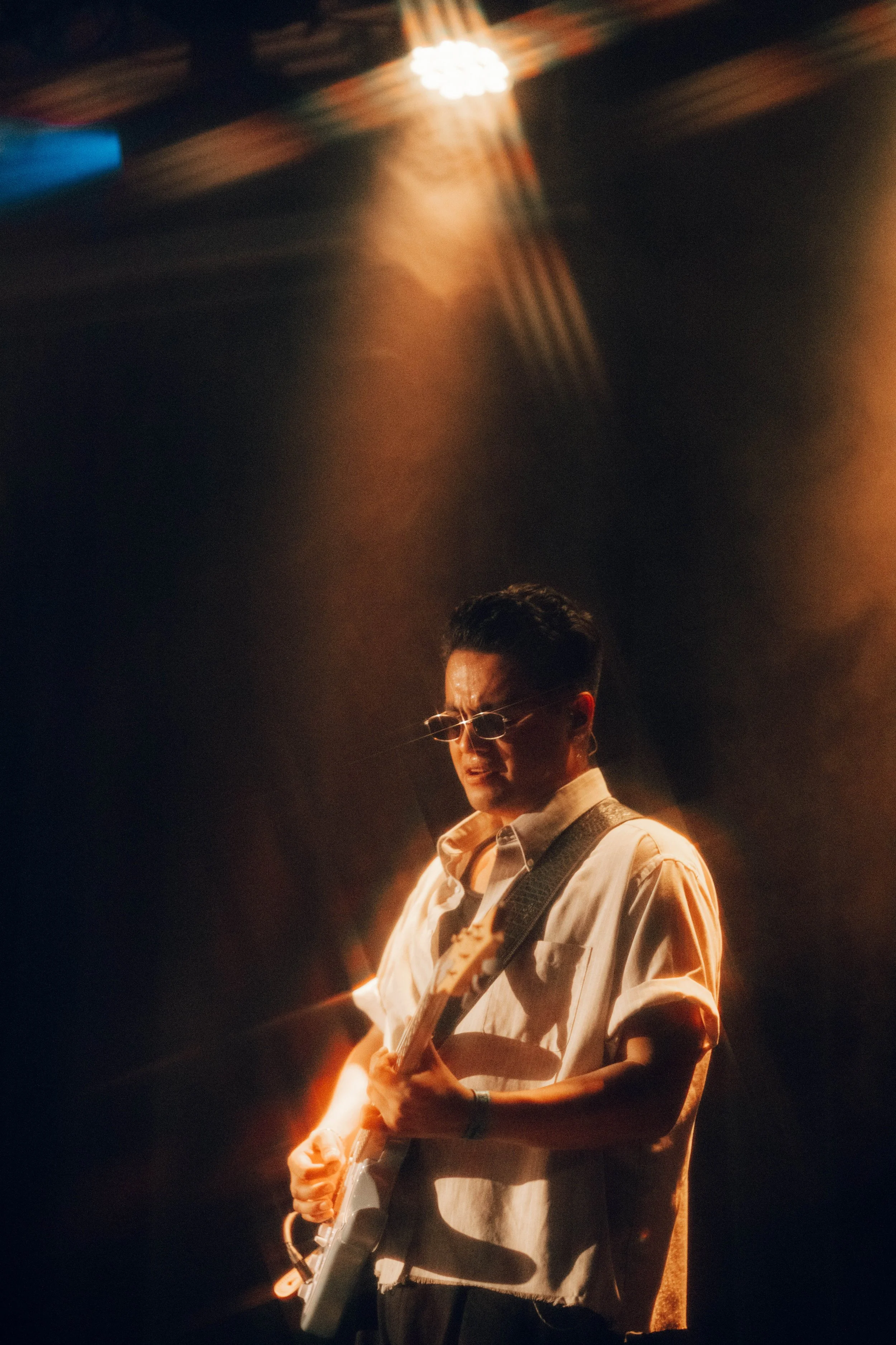 A man wearing glasses and a light-colored shirt playing an electric guitar on stage under warm lighting.