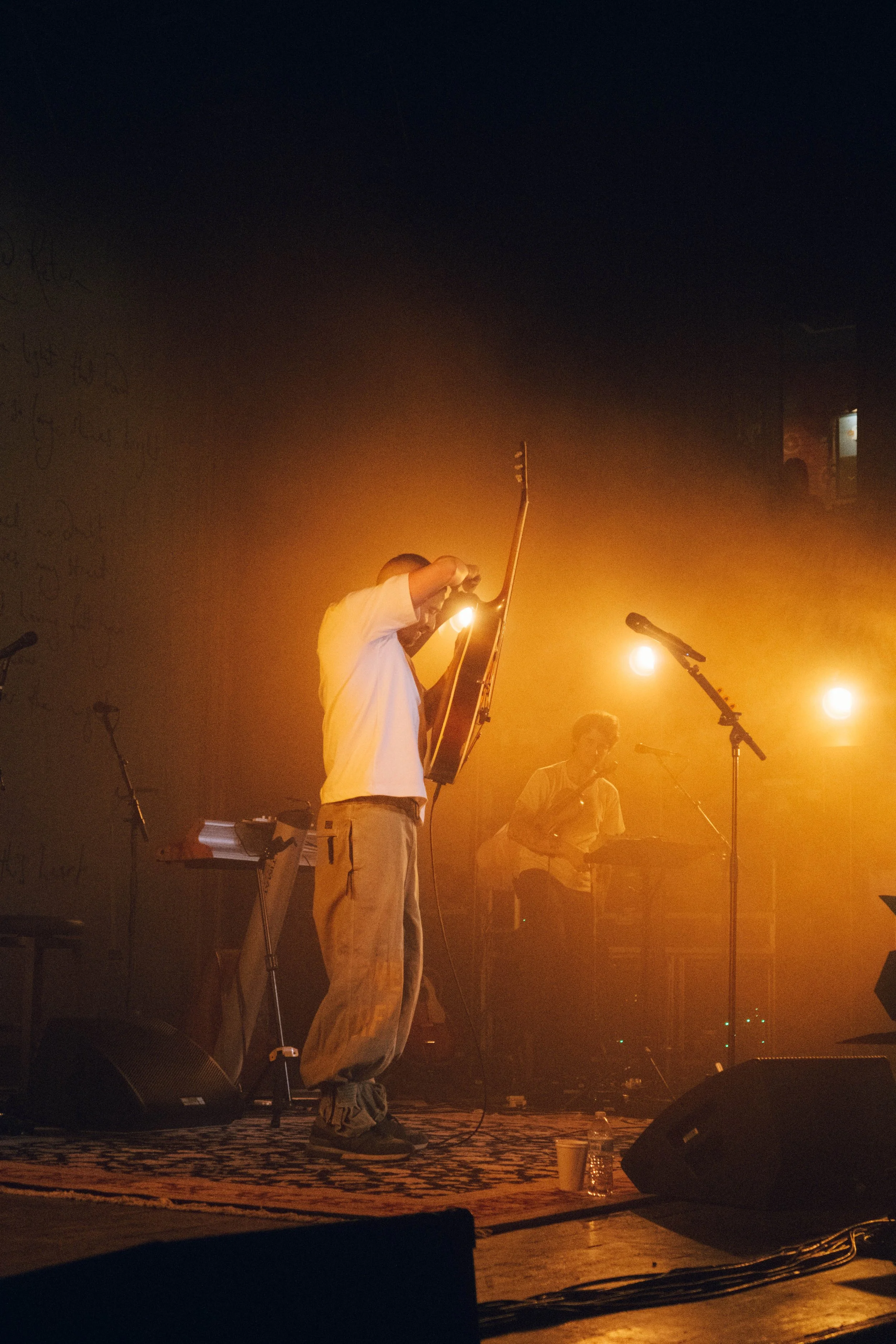 Dermot Kennedy performing on stage with a guitar, in a dimly lit venue with orange lighting, and another person in the background near a microphone at their concert in Chicago.