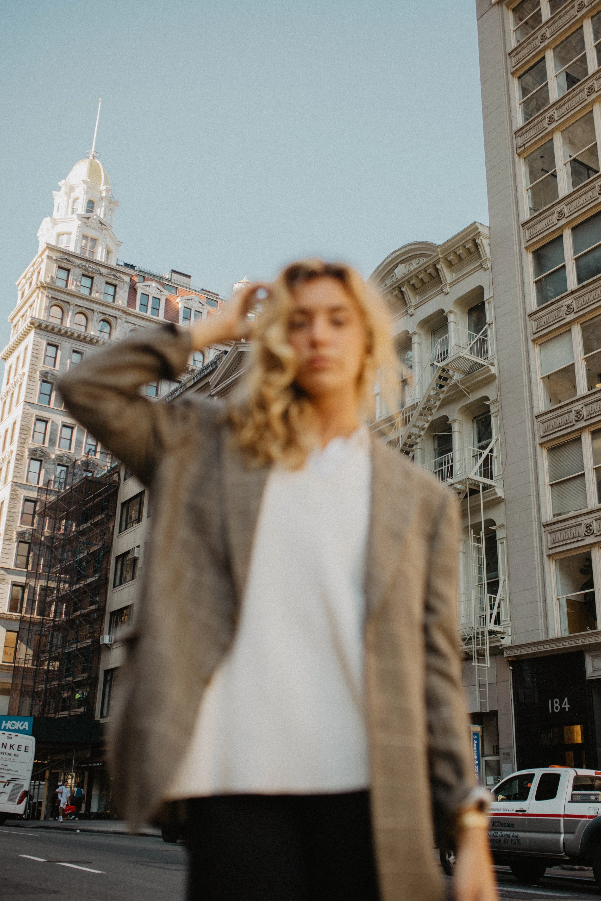 Blurred woman in business attire standing on city street with tall historic buildings and parked cars in background.