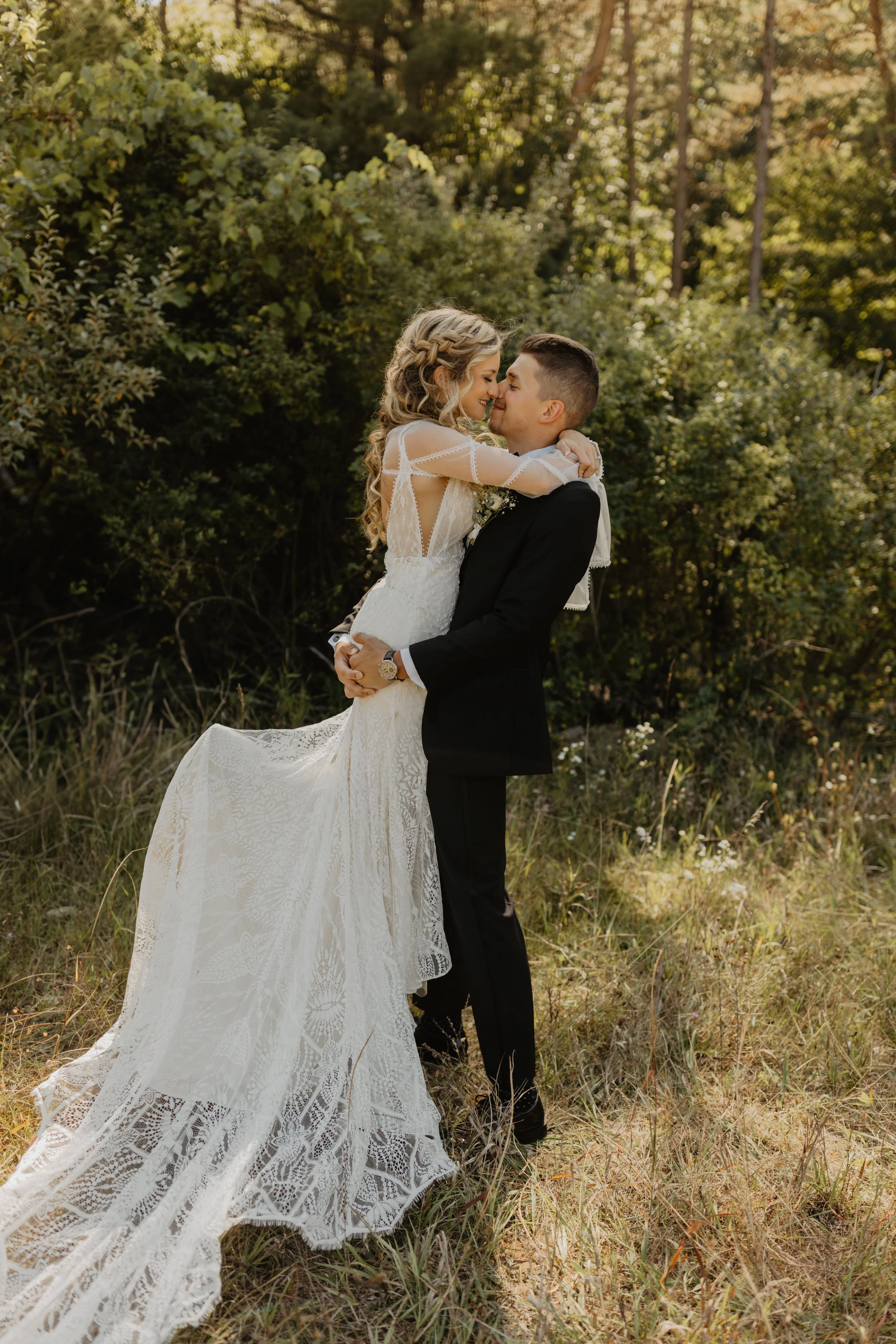 A bride and groom in wedding attire sharing a romantic moment outdoors, surrounded by greenery.