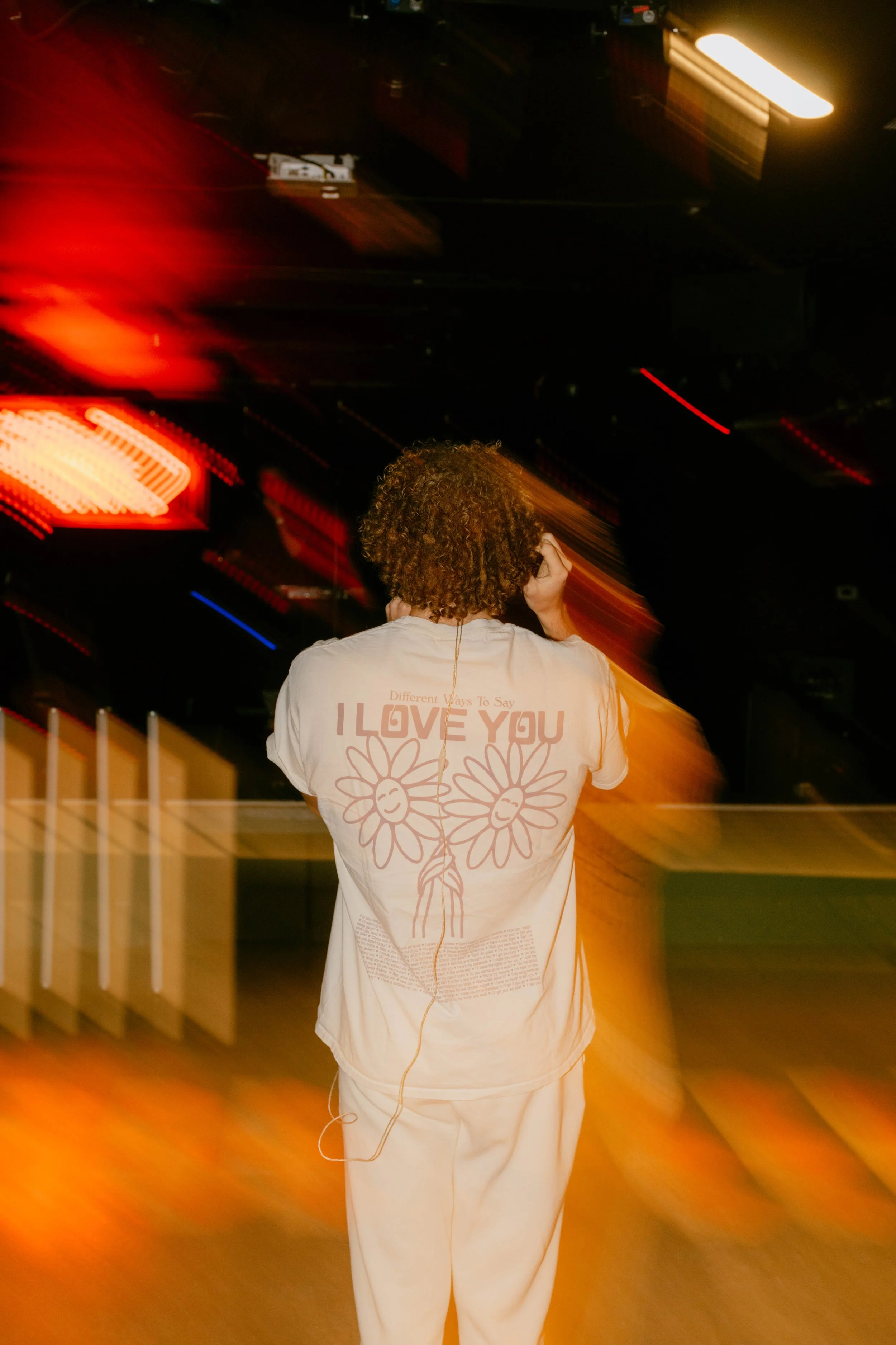 Person with curly hair wearing a white t-shirt with a floral design and the text "I LOVE YOU" on the back, standing at night with blurred colorful lights in the background.