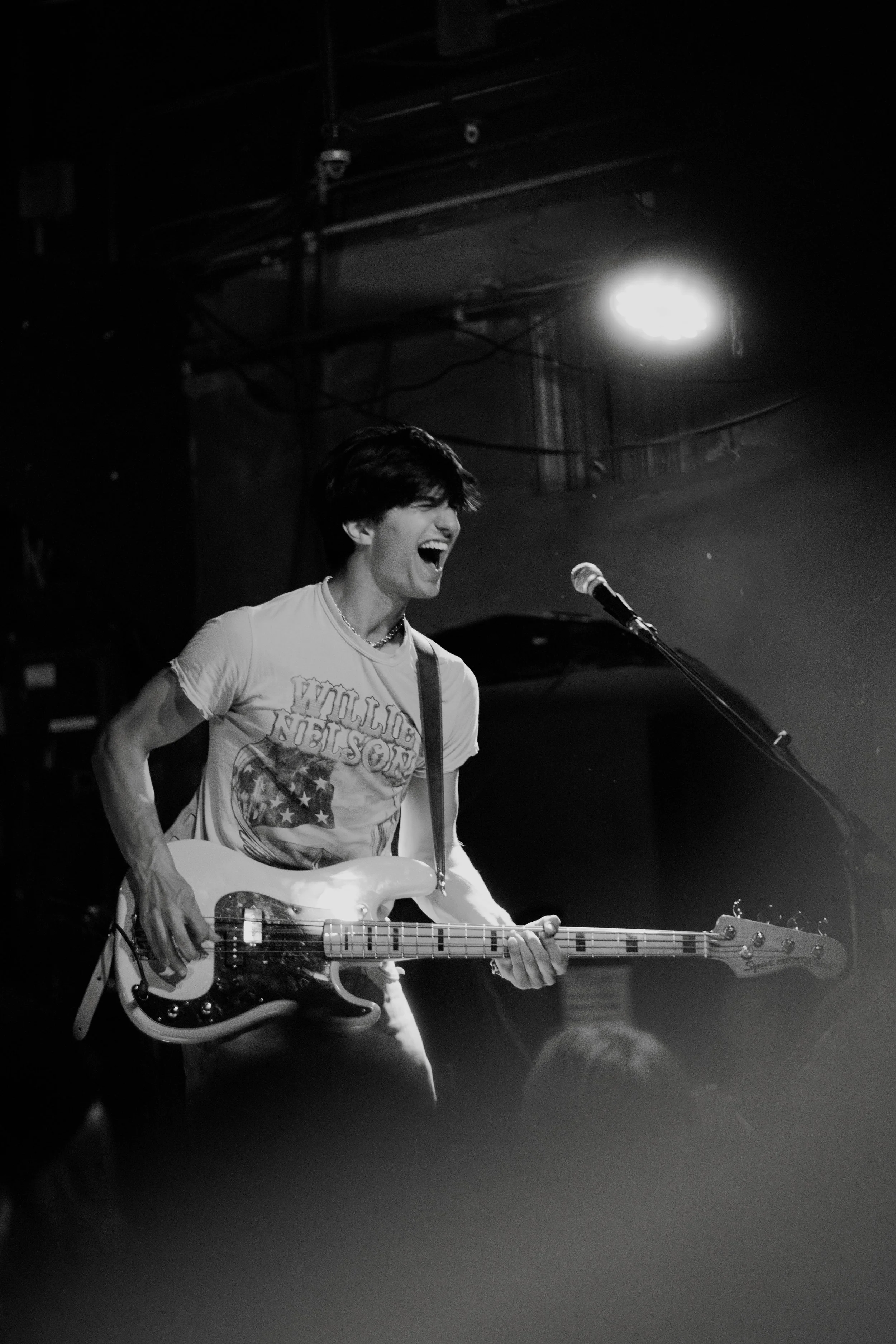 A young man playing a bass guitar on stage, smiling and singing into a microphone, in black and white.
