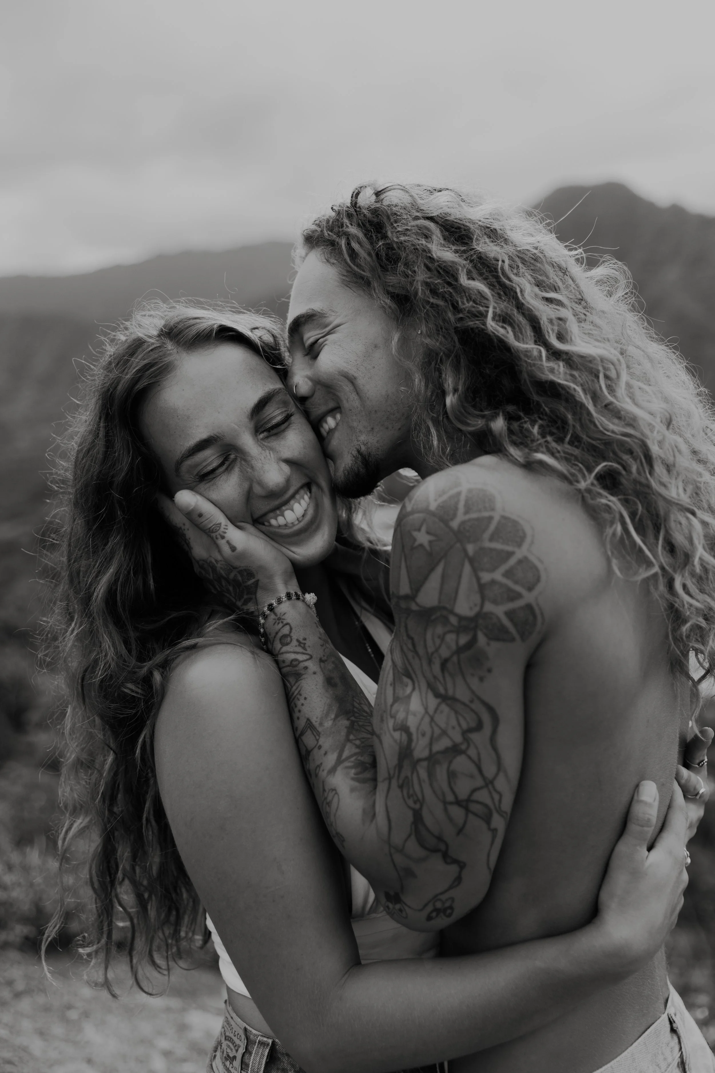A black and white photo of a couple hugging outdoors with mountains in the background, sharing a tender moment and smiling.
